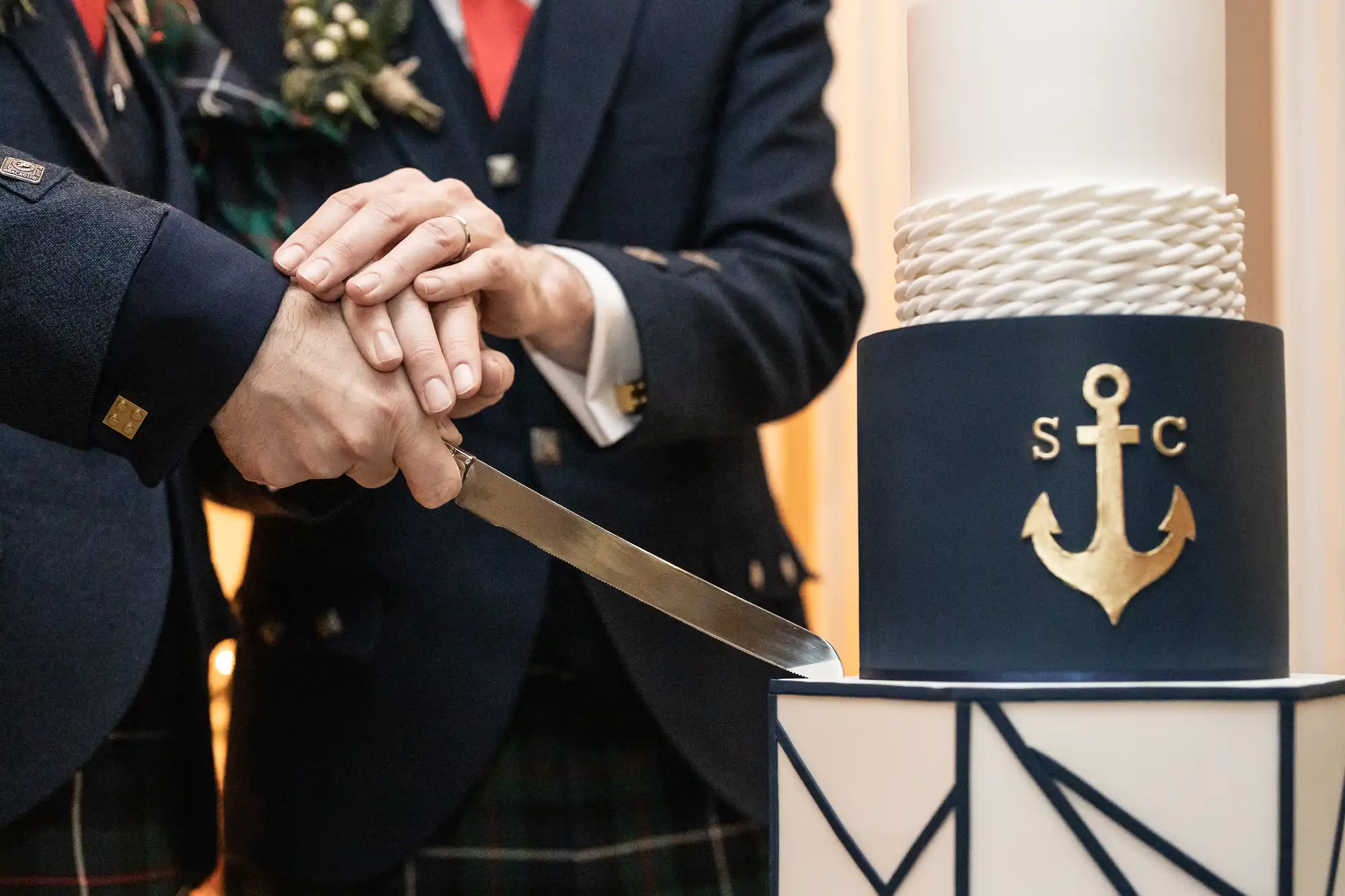 Two people in formal attire hold a knife to cut a multi-tiered navy blue and white cake adorned with an anchor and rope design with the initials "S" and "C".