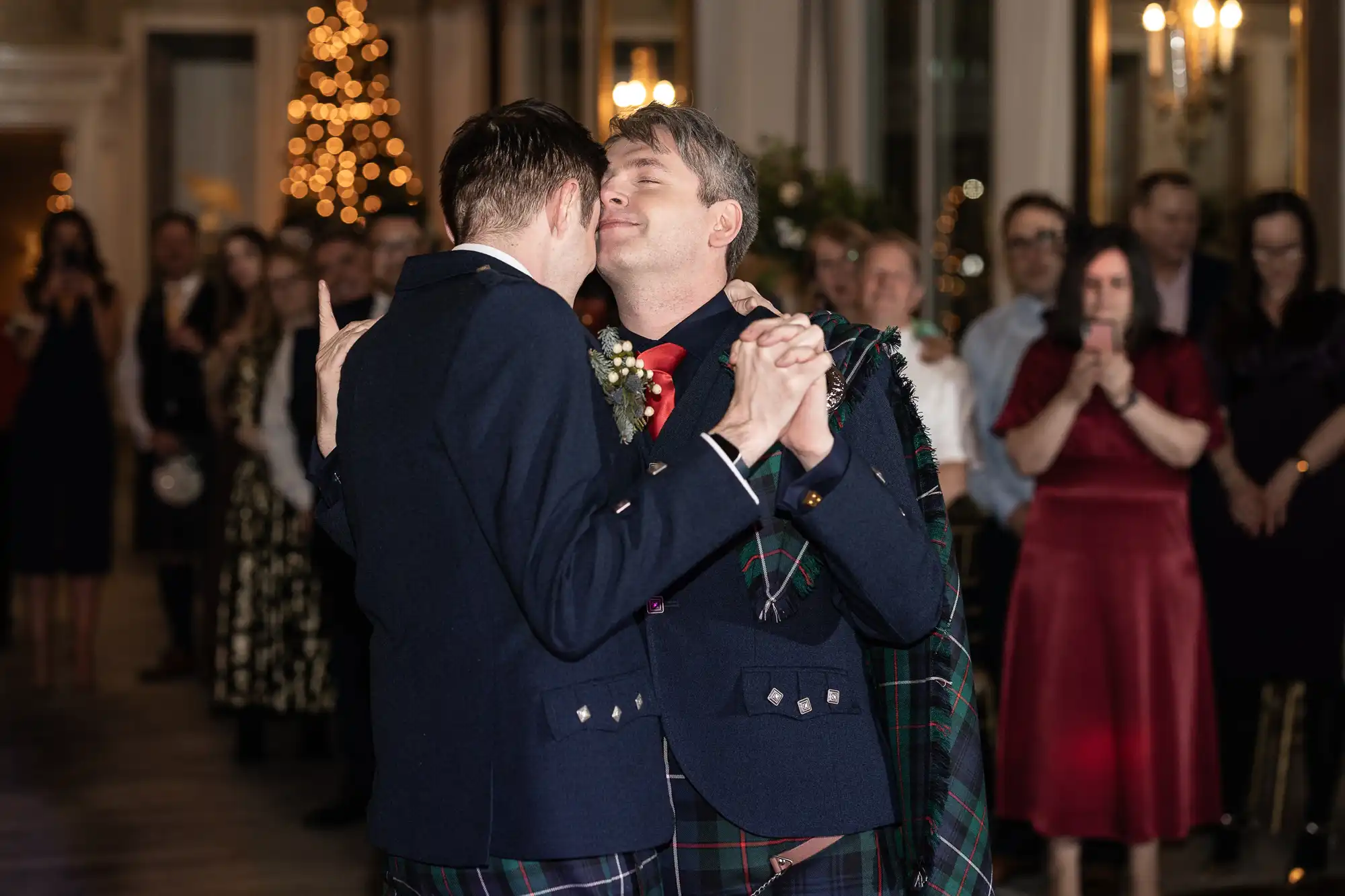 Two men dressed in suits and Scottish kilts share a dance at an indoor event with an audience watching in the background.