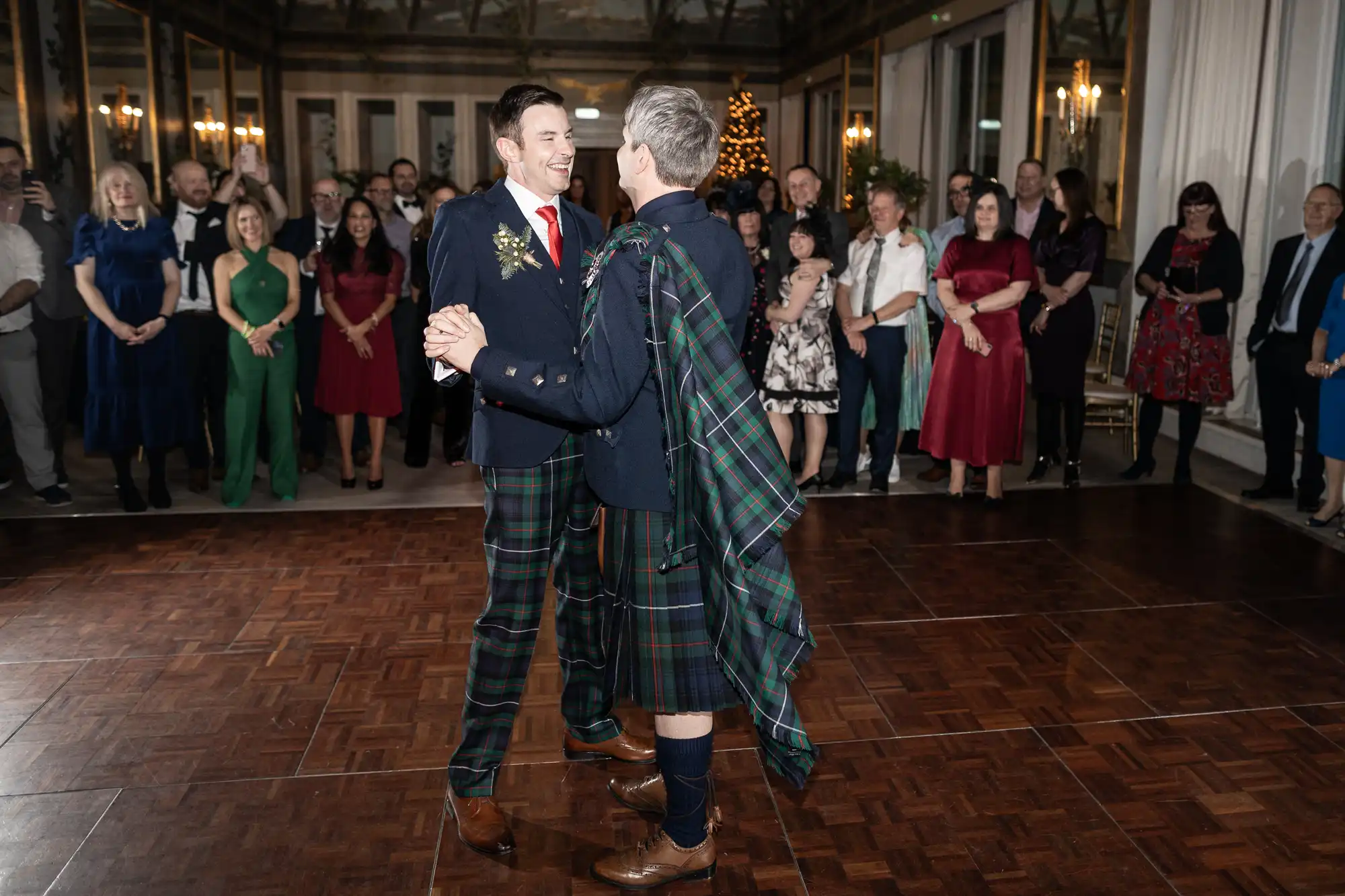Two men, both wearing tartan kilts, dance together in a large room with wooden floors, while a group of people watch and smile in the background.