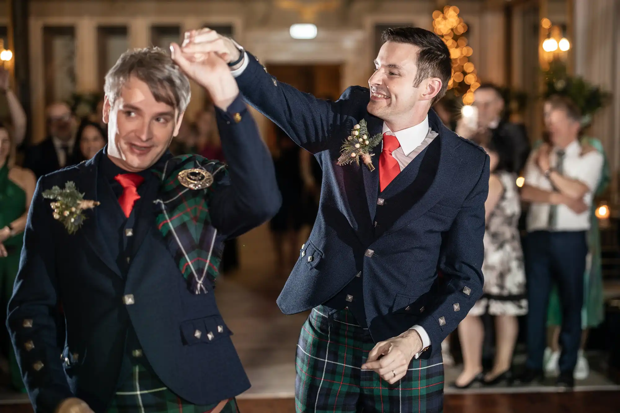 Two men dressed in formal Scottish attire dance together at an indoor event with a cheering crowd in the background.