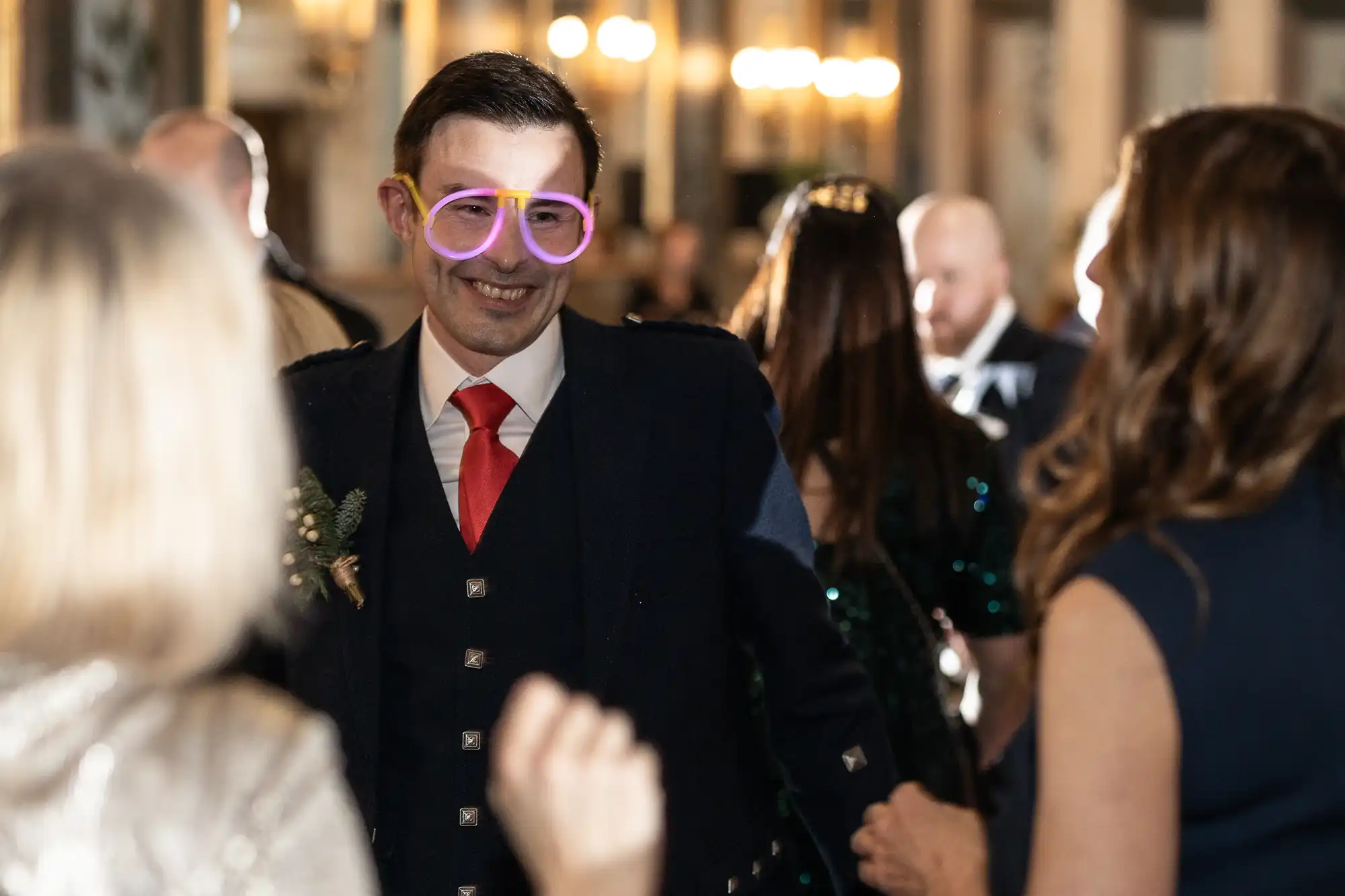 A man with neon pink glasses is smiling and interacting with guests at a formal event.