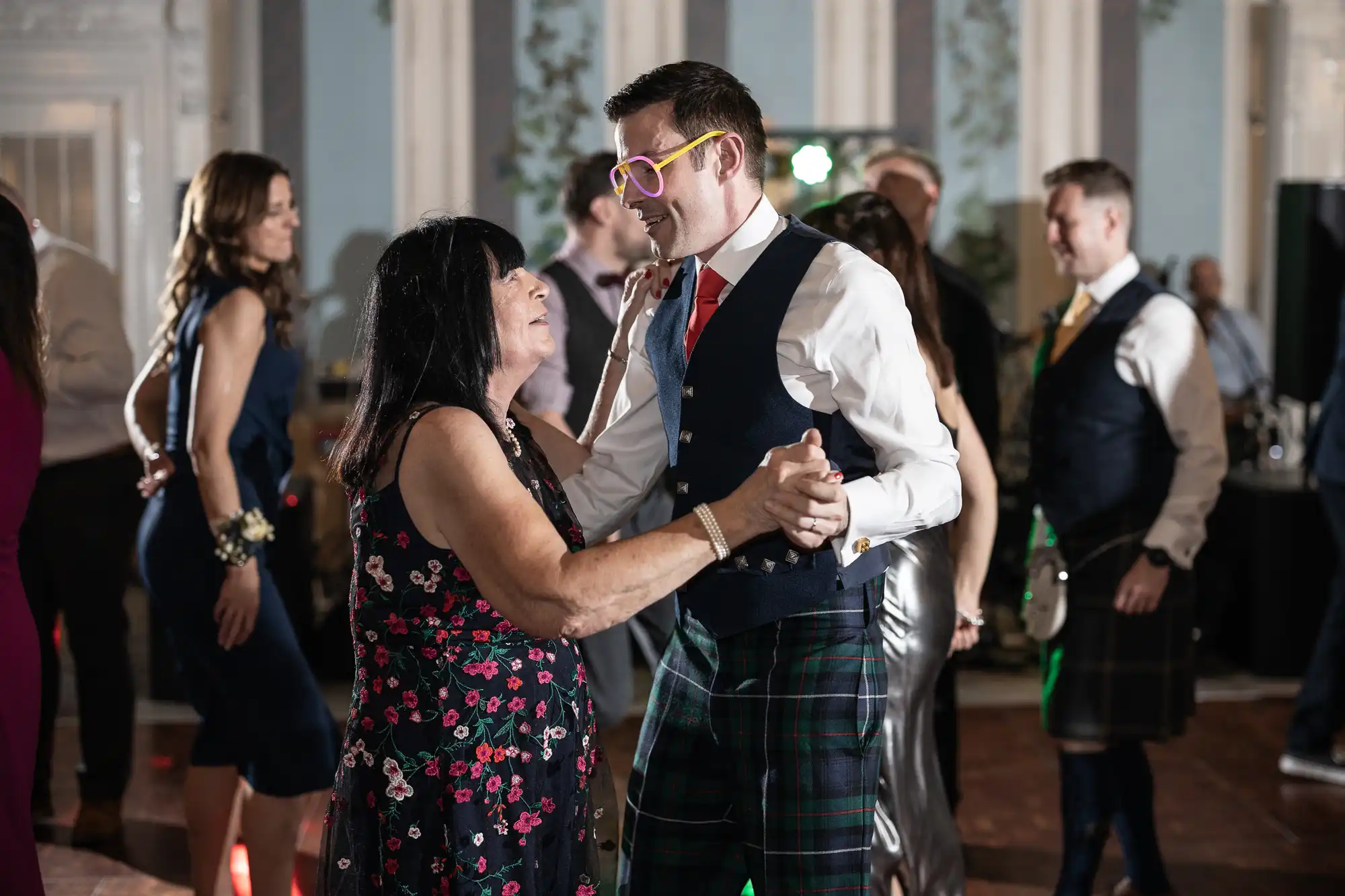 People dancing at an indoor event; a man in a vest and red tie wearing yellow glasses is dancing with a woman in a floral dress. Others are visible in the background.