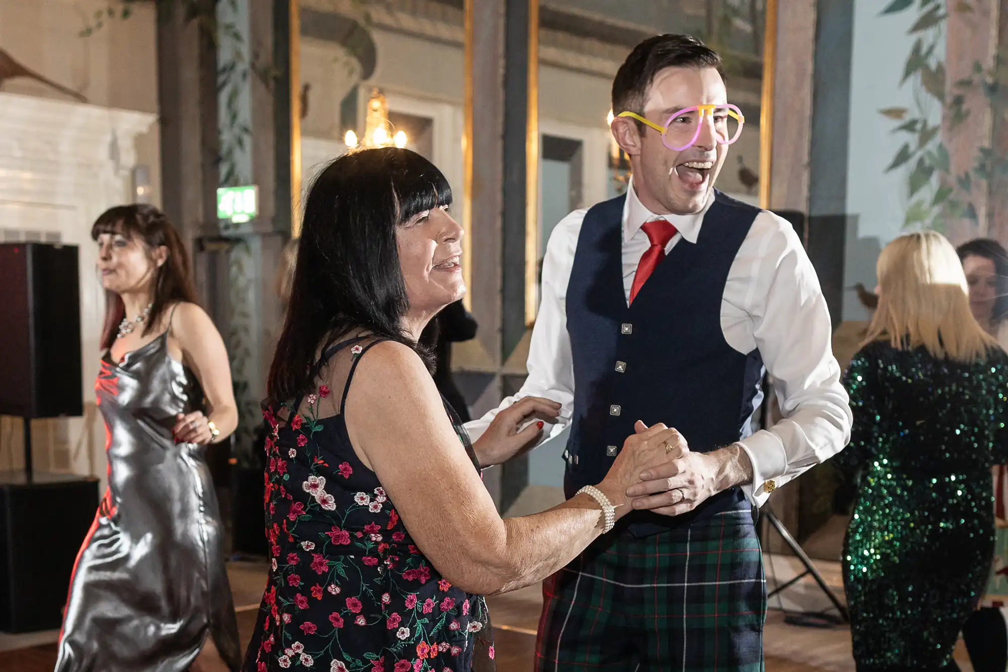 A man in a kilt and light-up glasses dances with a woman in a floral dress at an indoor event. Other people are dancing in the background.