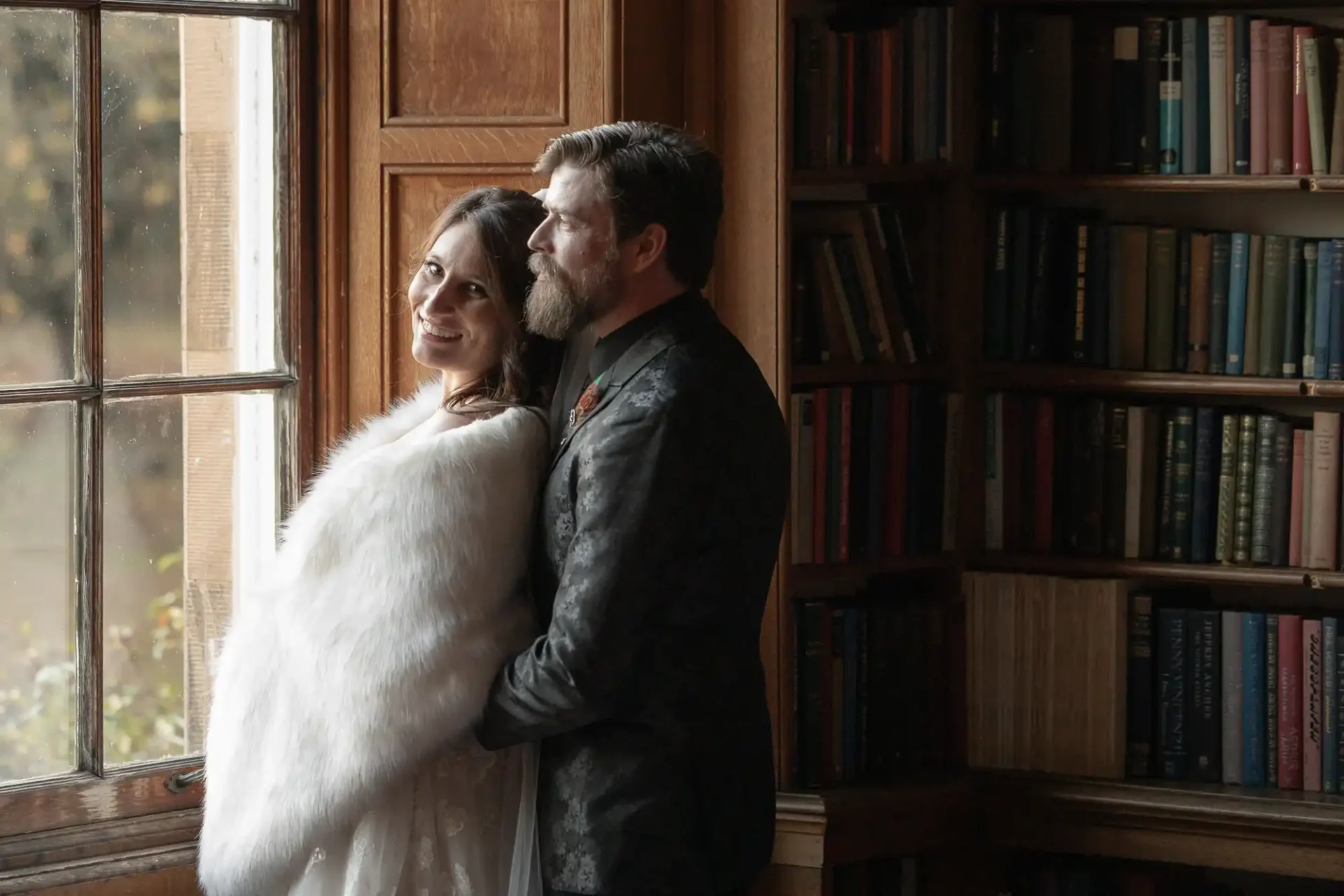 A couple stands by a window; the woman in a white shawl smiles at the camera while the man stands behind her, looking outside. Bookshelves fill the background.