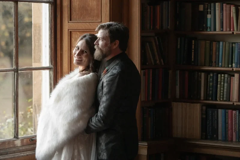 A couple photographed by Edinburgh wedding photographer, stands by a window; the woman in a white shawl smiles at the camera while the man stands behind her, looking outside. Bookshelves fill the background.
