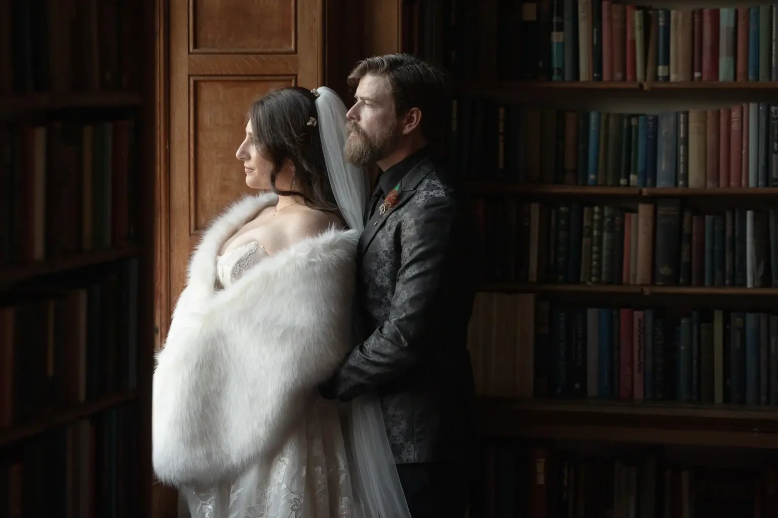 A bride and groom stand together in formal attire beside a bookshelf, gazing out of a window with soft natural light illuminating them.