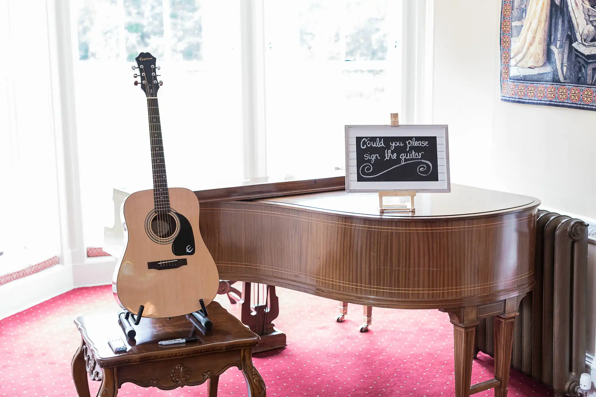 Acoustic guitar on a stand next to a grand piano with a sign saying "could you please sign the guitar" in a well-lit room with a painting on the wall.