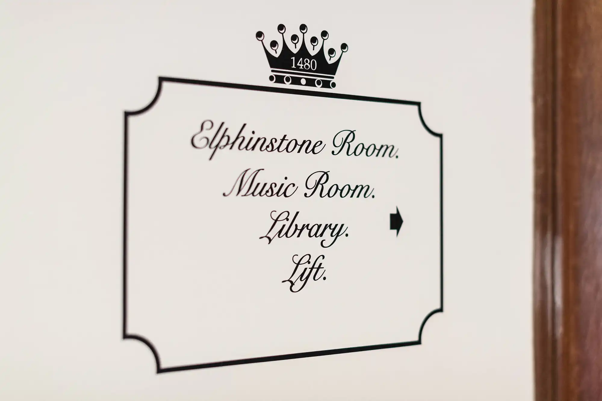 Sign on a wall indicating directions to "elphinstone room," "music room," "library," and "lift," featuring ornate calligraphy and a crown symbol.