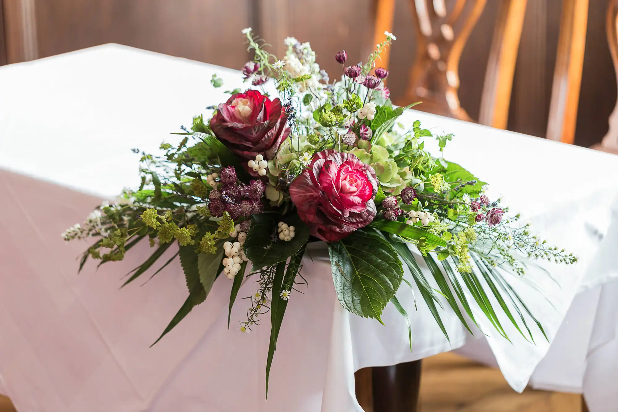 Elegant floral centerpiece with deep red roses and assorted greenery on a white draped table, in a warmly lit indoor setting.