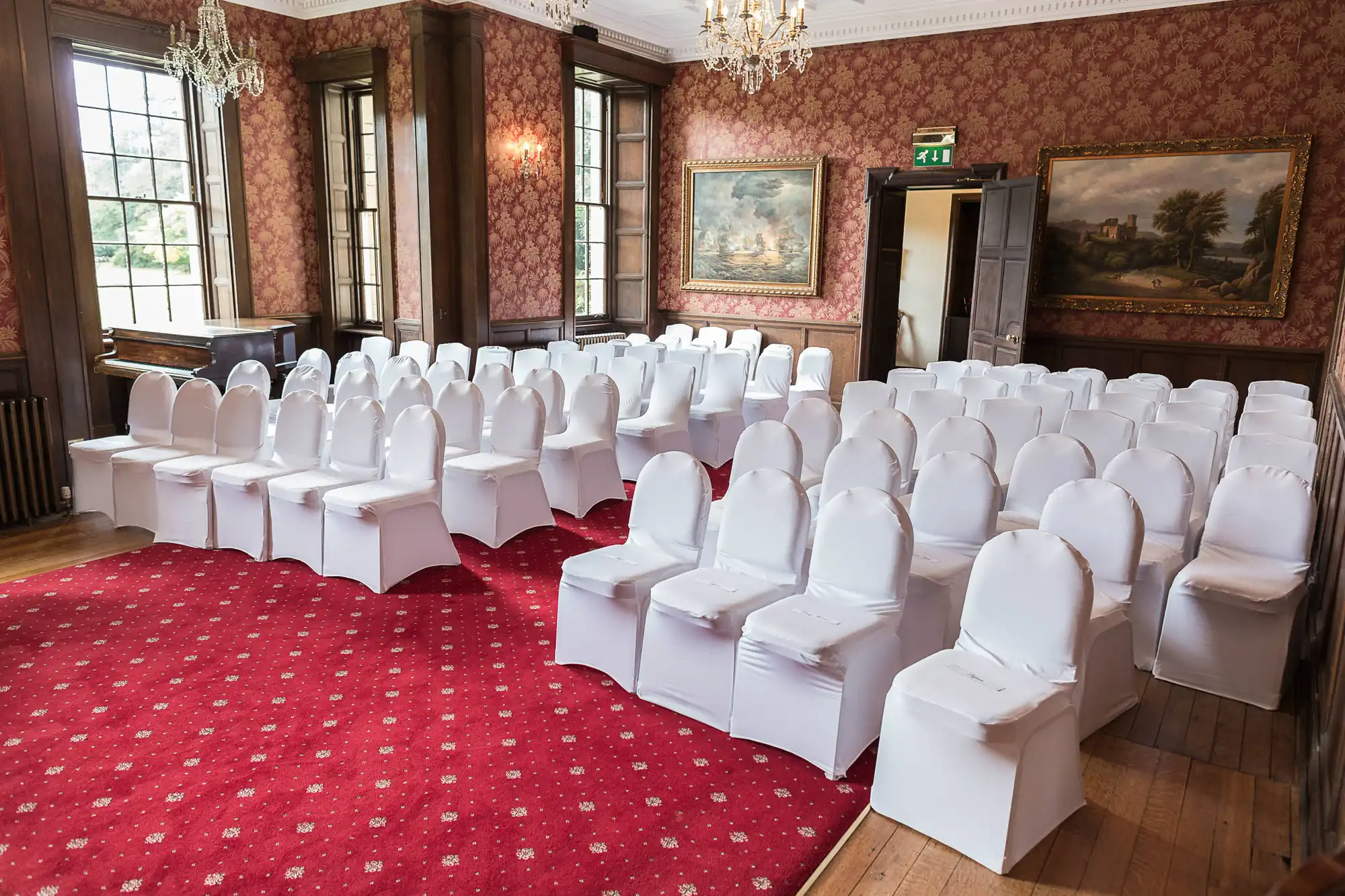 Elegant event room with rows of white chairs arranged on a red carpet, large windows, and classic paintings on the walls.