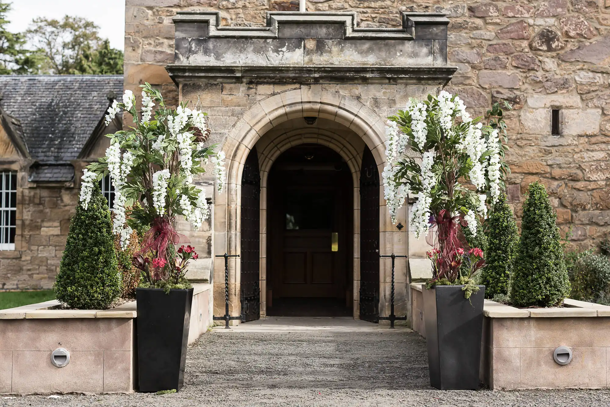 Arched stone entrance of an historic building flanked by ornate planters with white flowers and greenery.