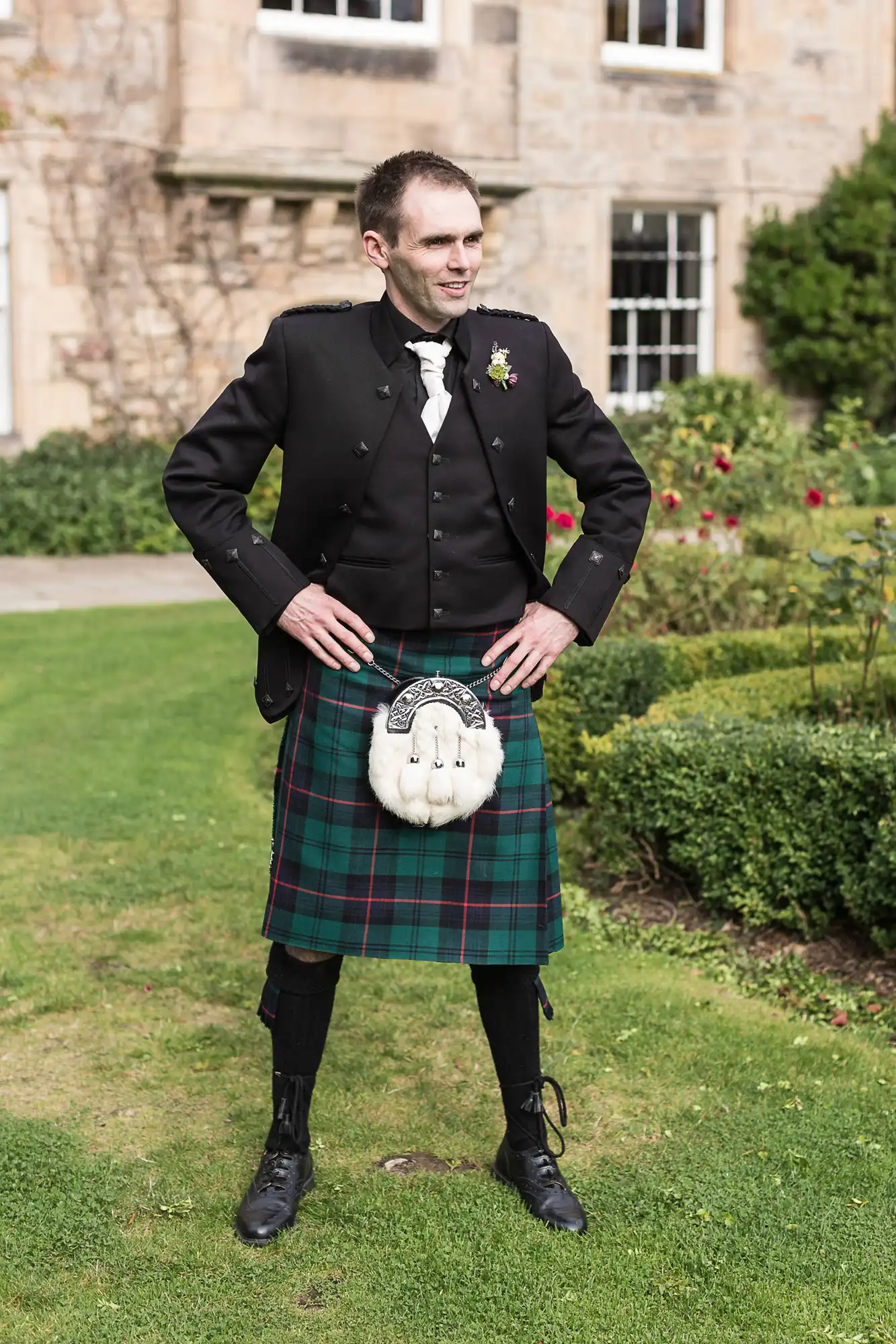 Man in traditional scottish attire, including kilt and sporran, standing outdoors with a castle-like building and gardens in the background.