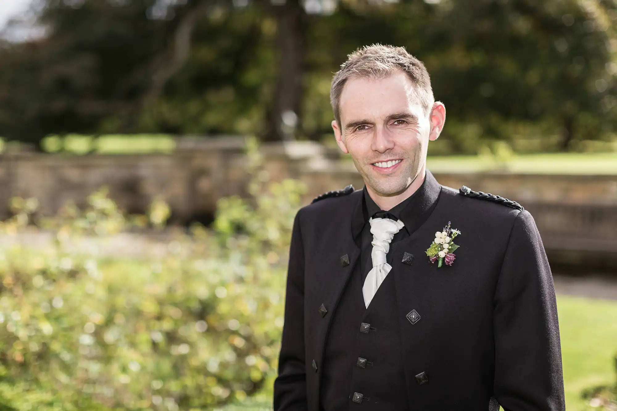 A man in a formal kilt jacket and tie smiles at the camera, outdoors with greenery and sunlight in the background.