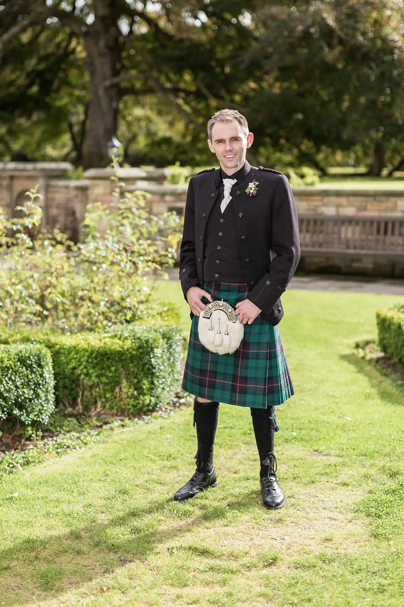 A man in a traditional scottish kilt and jacket smiling in a garden setting, holding a sporran.