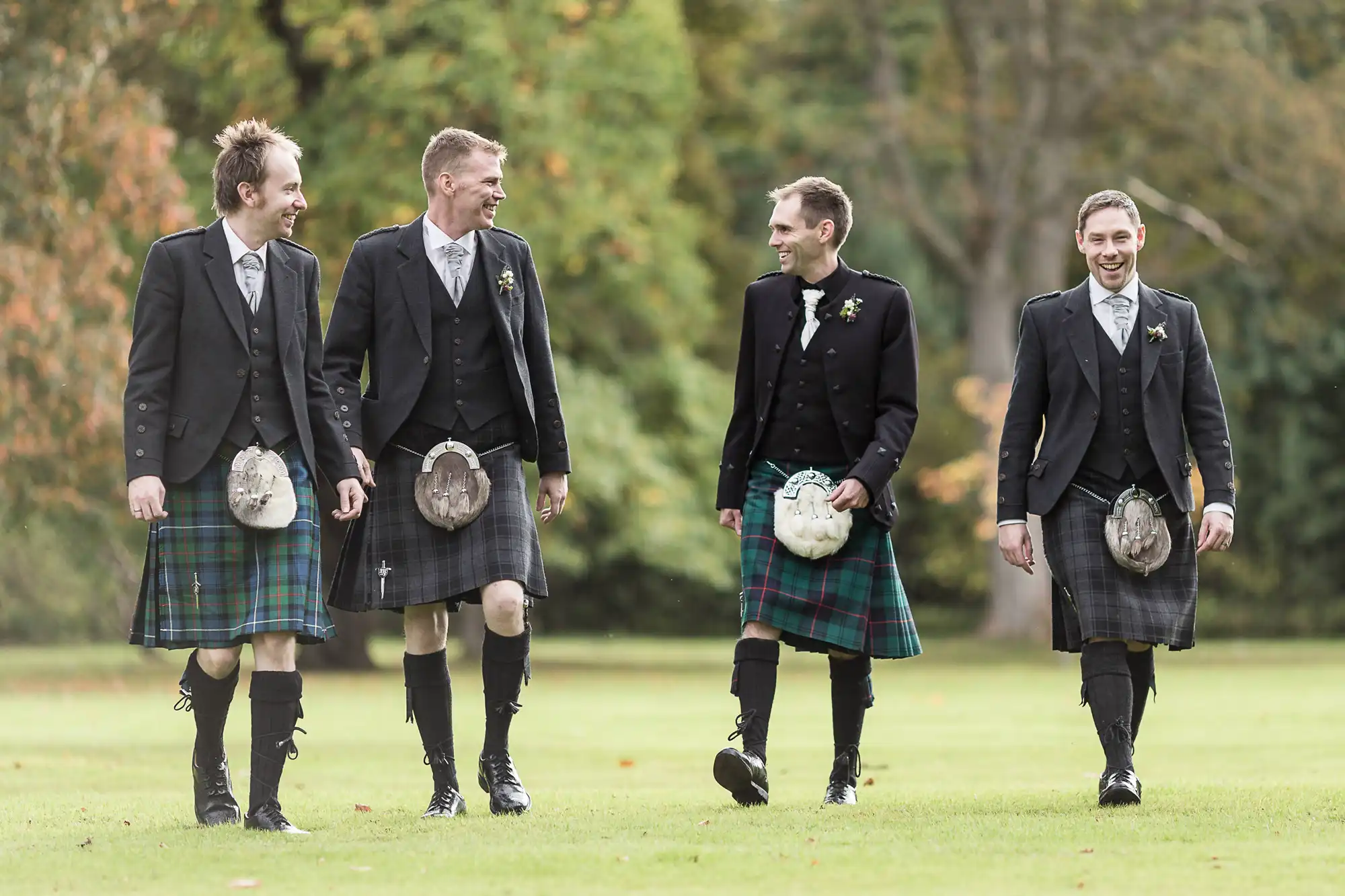 Four men wearing traditional scottish kilts, laughing and walking together on a grassy field.