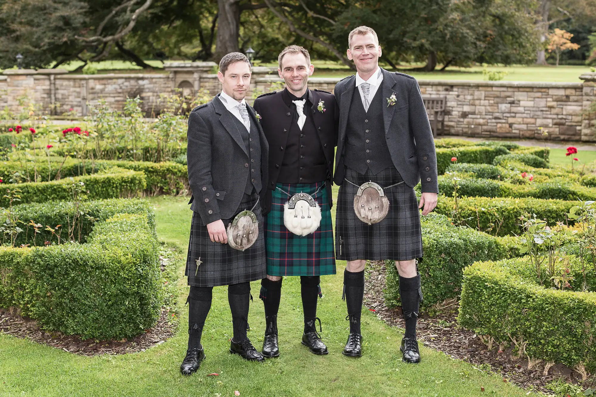Three men wearing traditional scottish kilts and sporran pouches posing together in a garden.