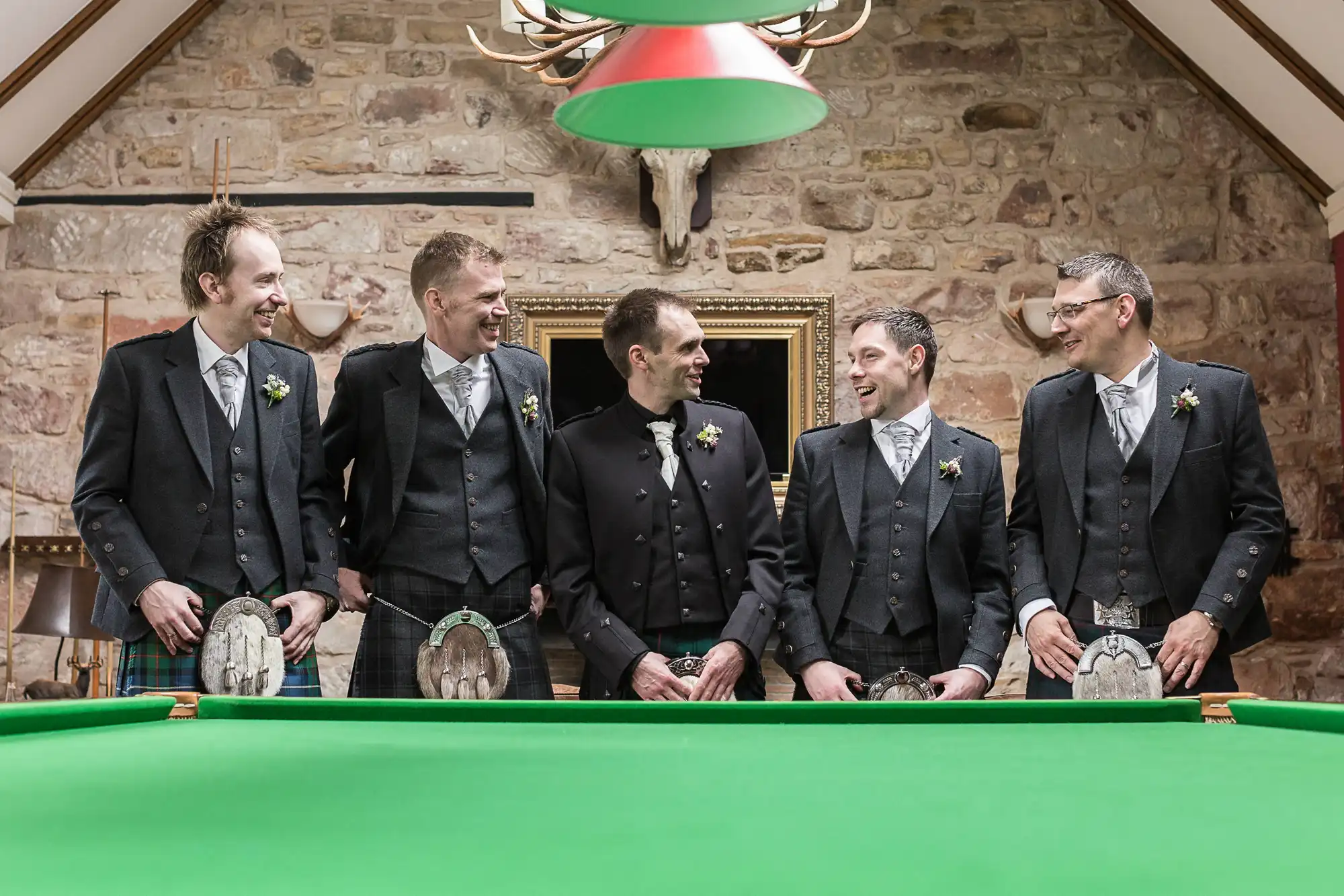 Five men in suits and kilts standing and laughing around a green billiards table in a rustic room with a stone wall backdrop.