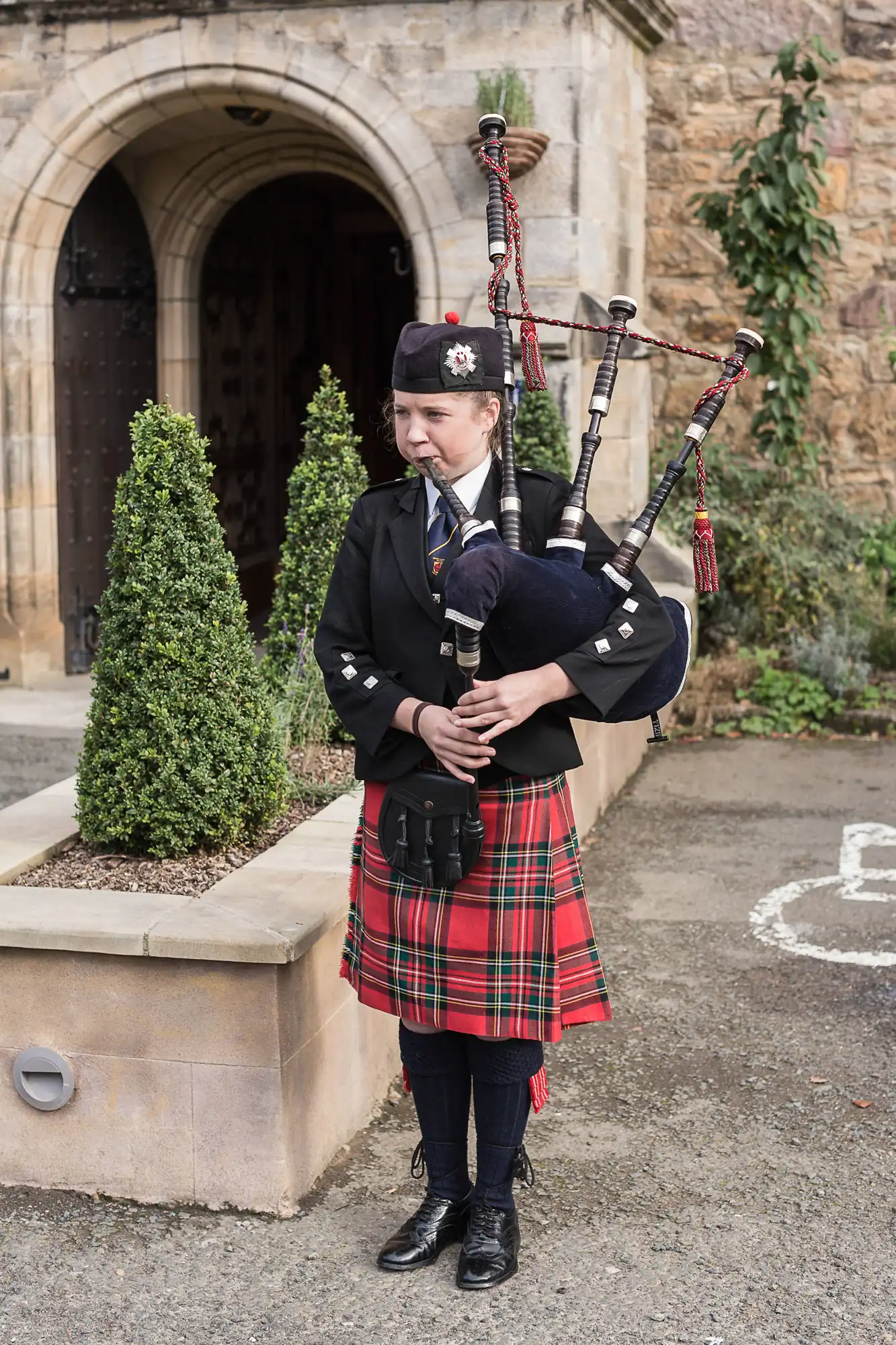 A woman in traditional scottish attire, including a tartan kilt, playing the bagpipes outside a stone church entrance.
