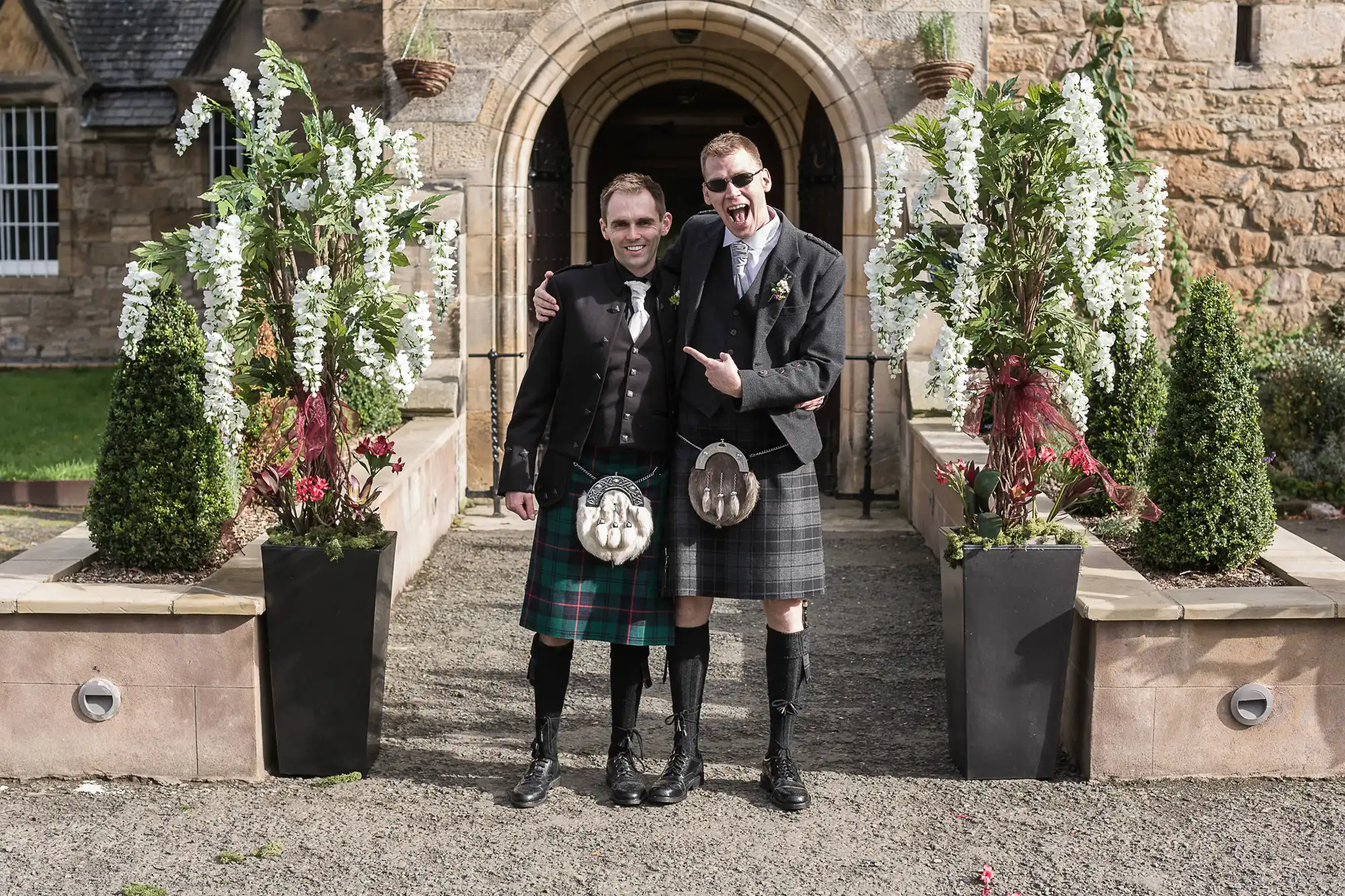 Two men in traditional scottish kilts smiling and standing arm in arm in front of a stone building with floral decorations.