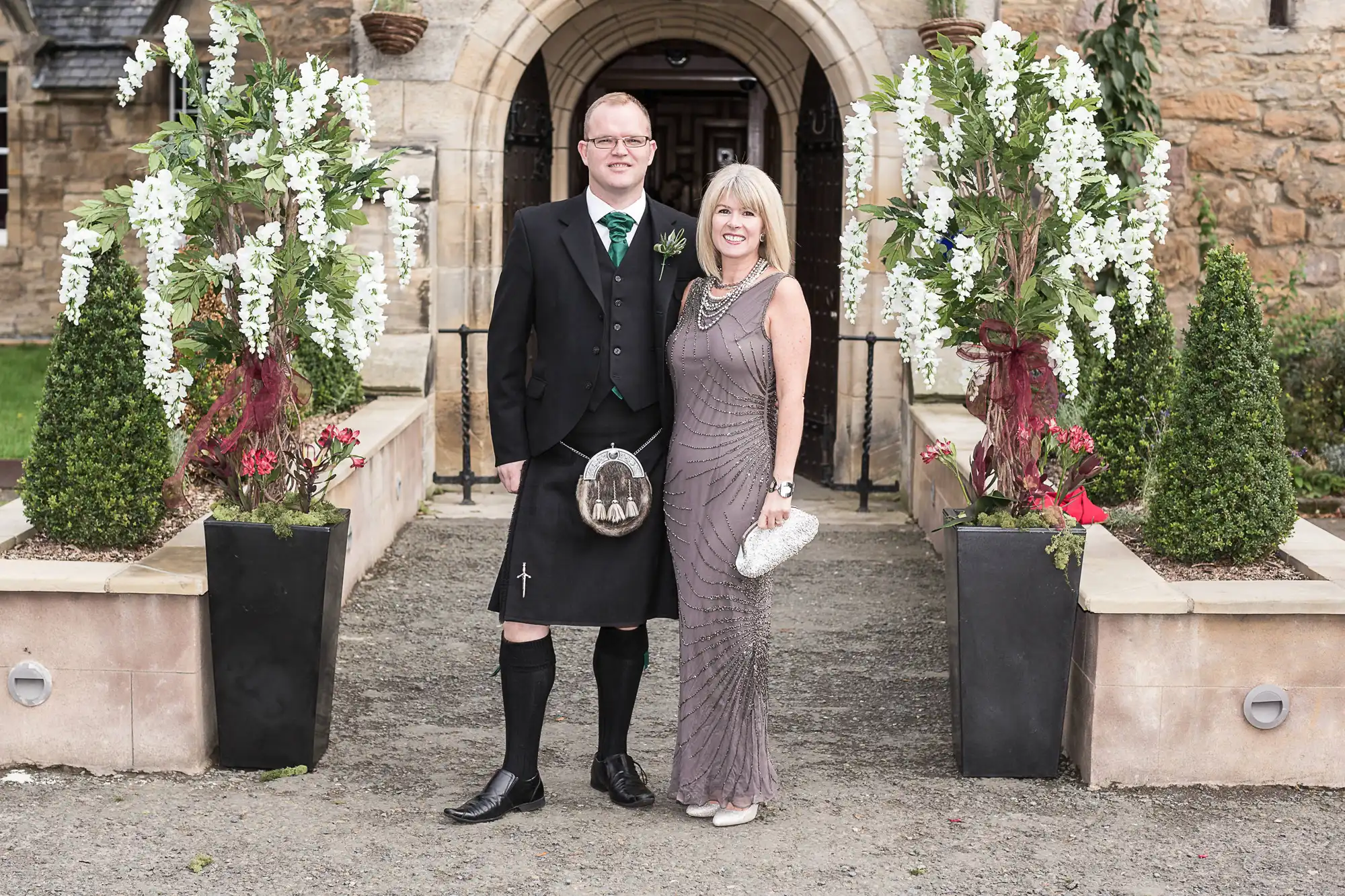 A couple dressed formally, the man in a kilt and the woman in a long taupe dress, standing in front of a church entrance, both smiling.