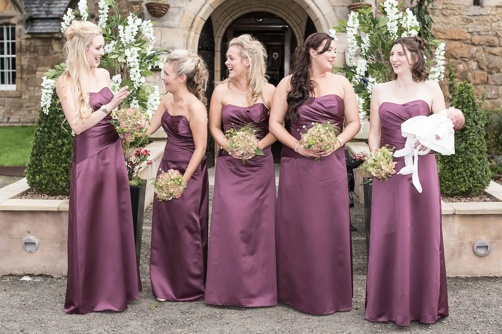 Five women in matching burgundy dresses holding bouquets, with one holding a baby, standing outside a stone building.