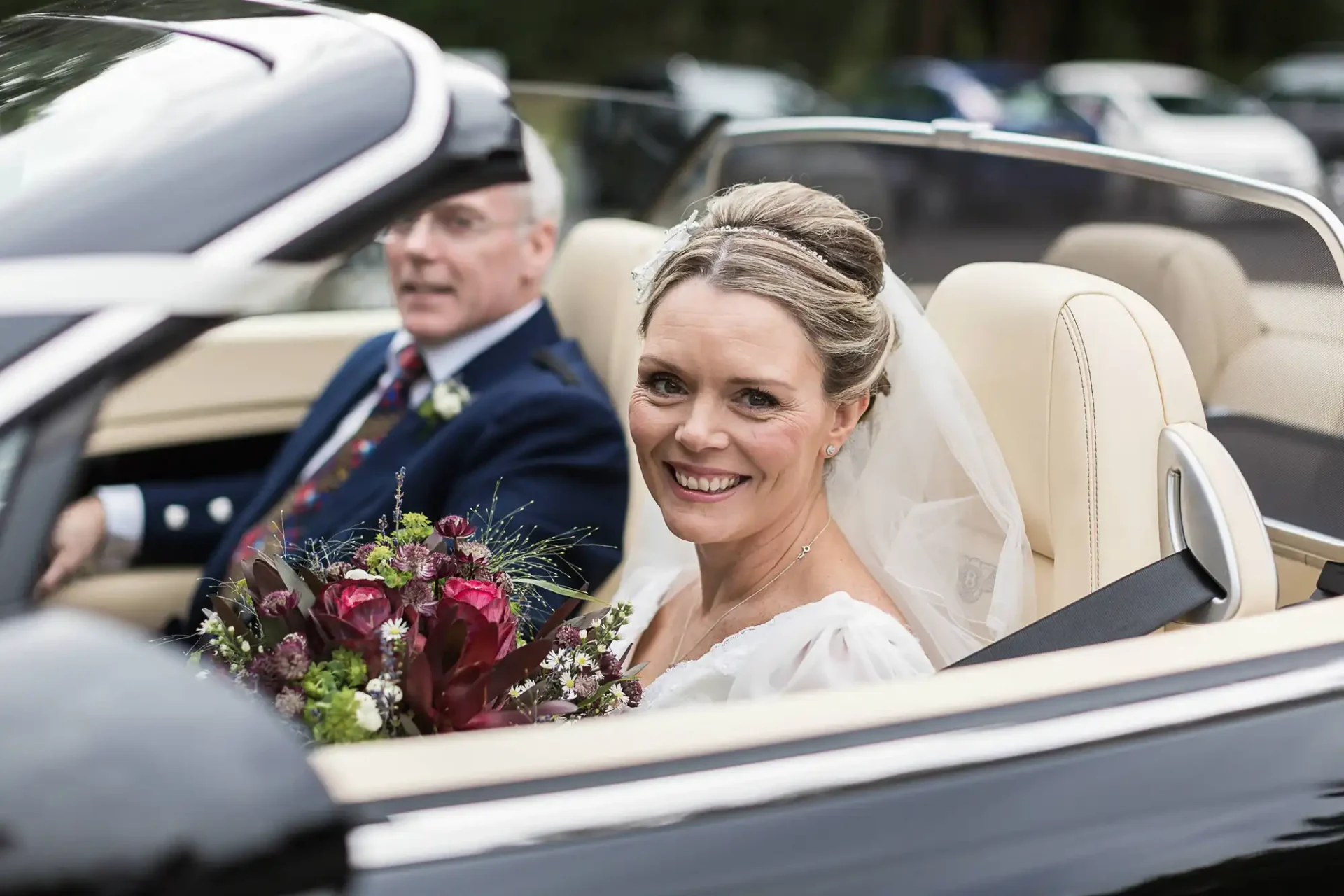 Carberry Tower wedding photos: A bride smiling in a convertible car with an older man driving, both dressed formally for a wedding, surrounded by trees.