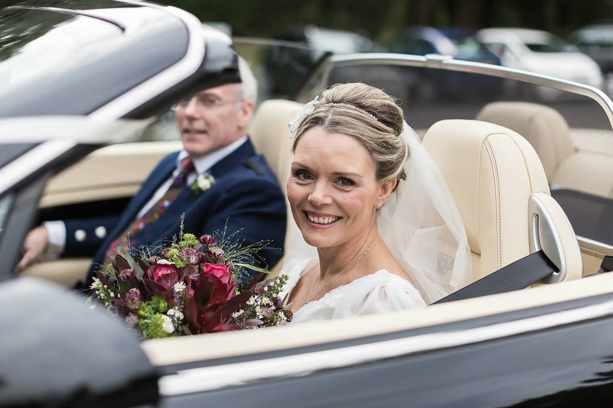 A bride smiling in a convertible car with an older man driving, both dressed formally for a wedding, surrounded by trees.