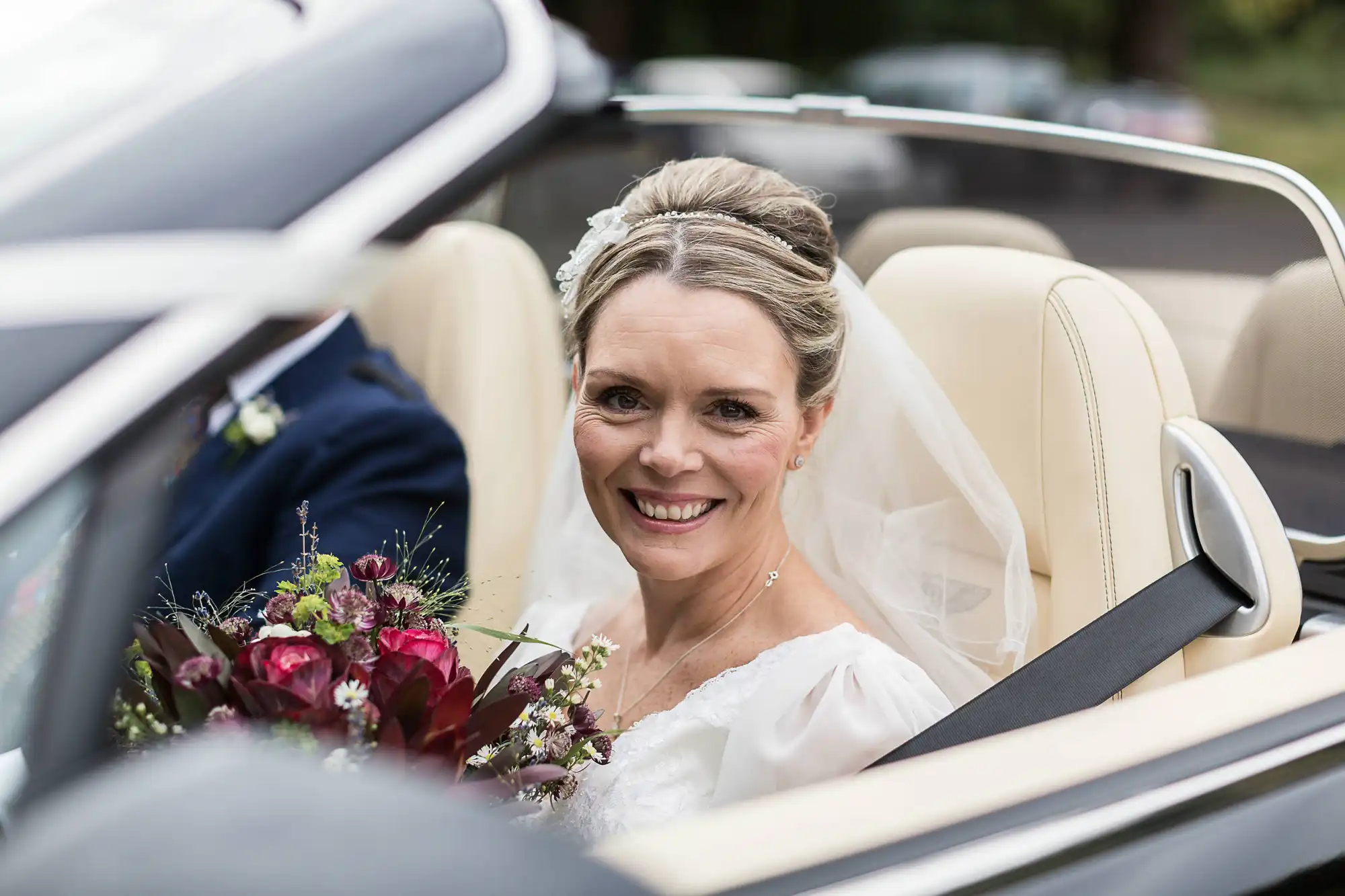 A smiling bride in a white dress and veil sits in a convertible car, holding a bouquet of flowers, ready to depart.