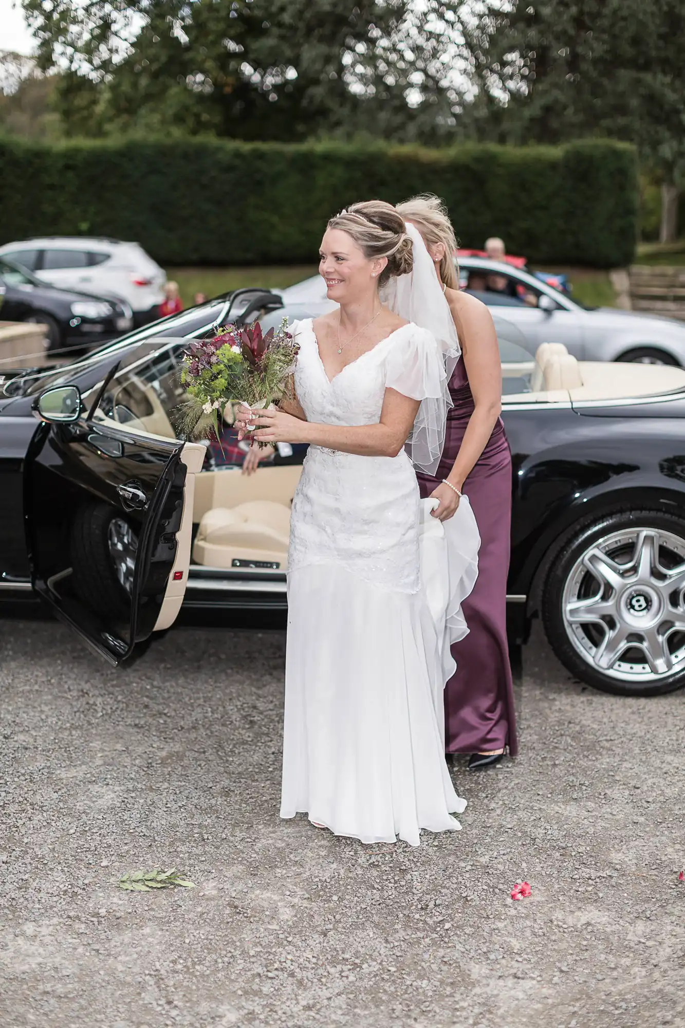 A bride in a white dress holding a bouquet smiles as she stands beside a luxury car, assisted by a bridesmaid in purple.