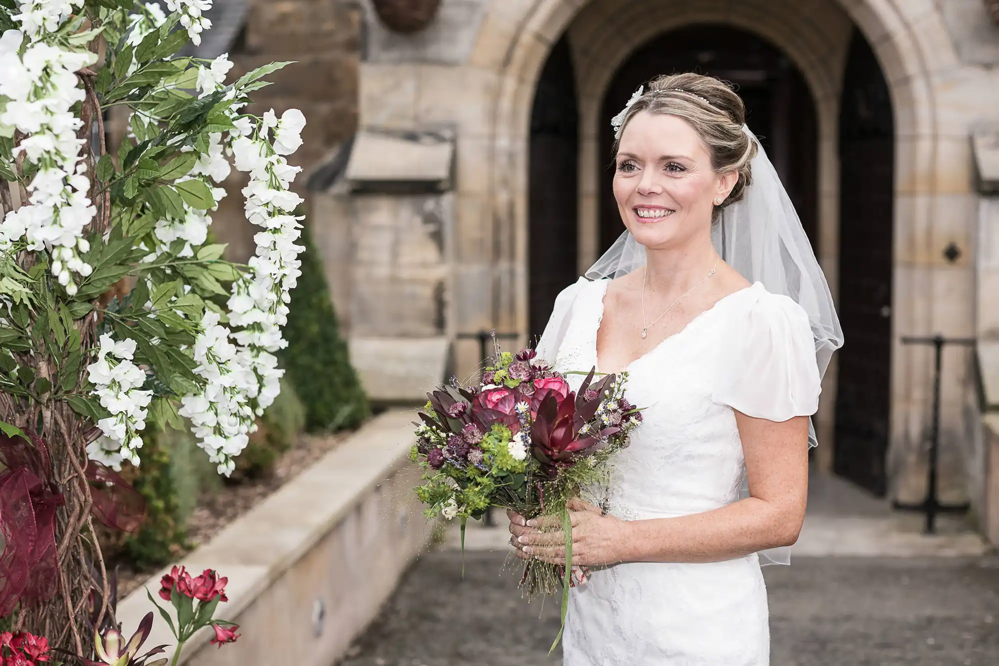 A smiling bride holding a bouquet, standing in front of a stone building with white flowers on the left.