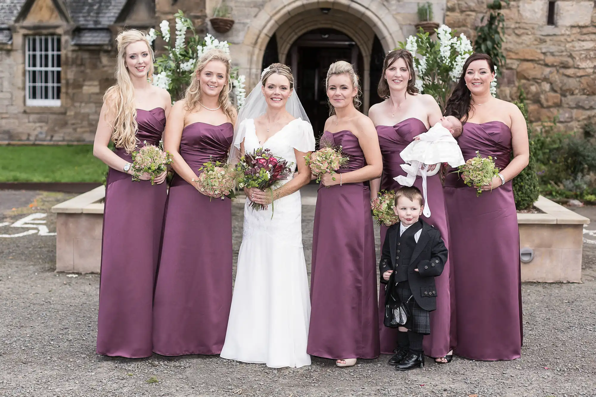 A bride in white stands with five bridesmaids in purple dresses and a young boy in a suit outside a stone building.