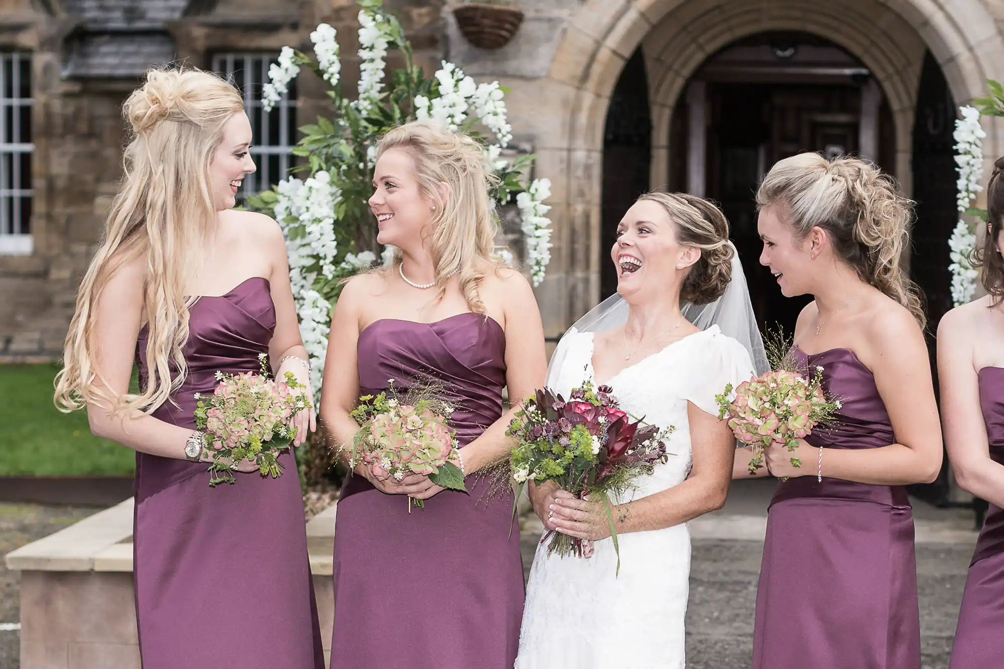 A bride in a white dress laughing joyfully with four bridesmaids in burgundy dresses, holding bouquets, in front of an old stone building.