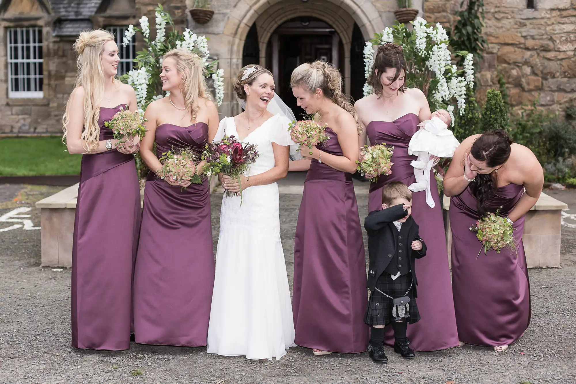 Five bridesmaids in purple dresses and a bride in white laugh together, while a young boy in a kilt peeks through one bridesmaid's legs.