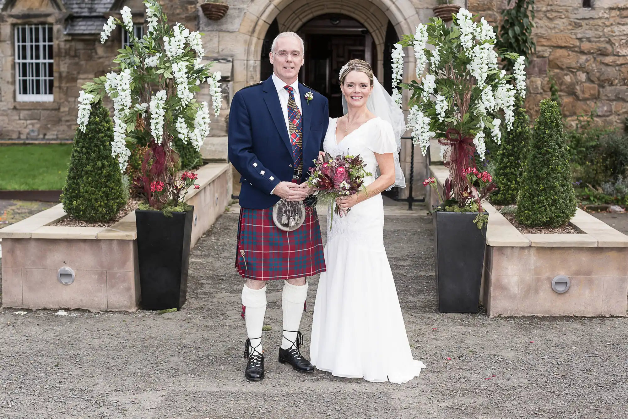 A bride and groom smiling in front of a stone building, with the groom wearing traditional scottish attire and the bride in a white dress, holding a bouquet.
