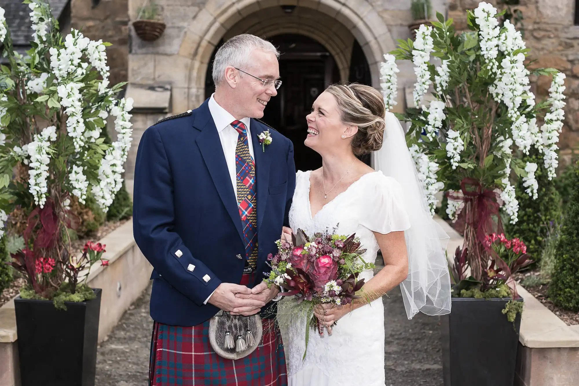 A bride and groom smiling at each other, the groom in a kilt and the bride in a white dress, standing at a church entrance decorated with white flowers.