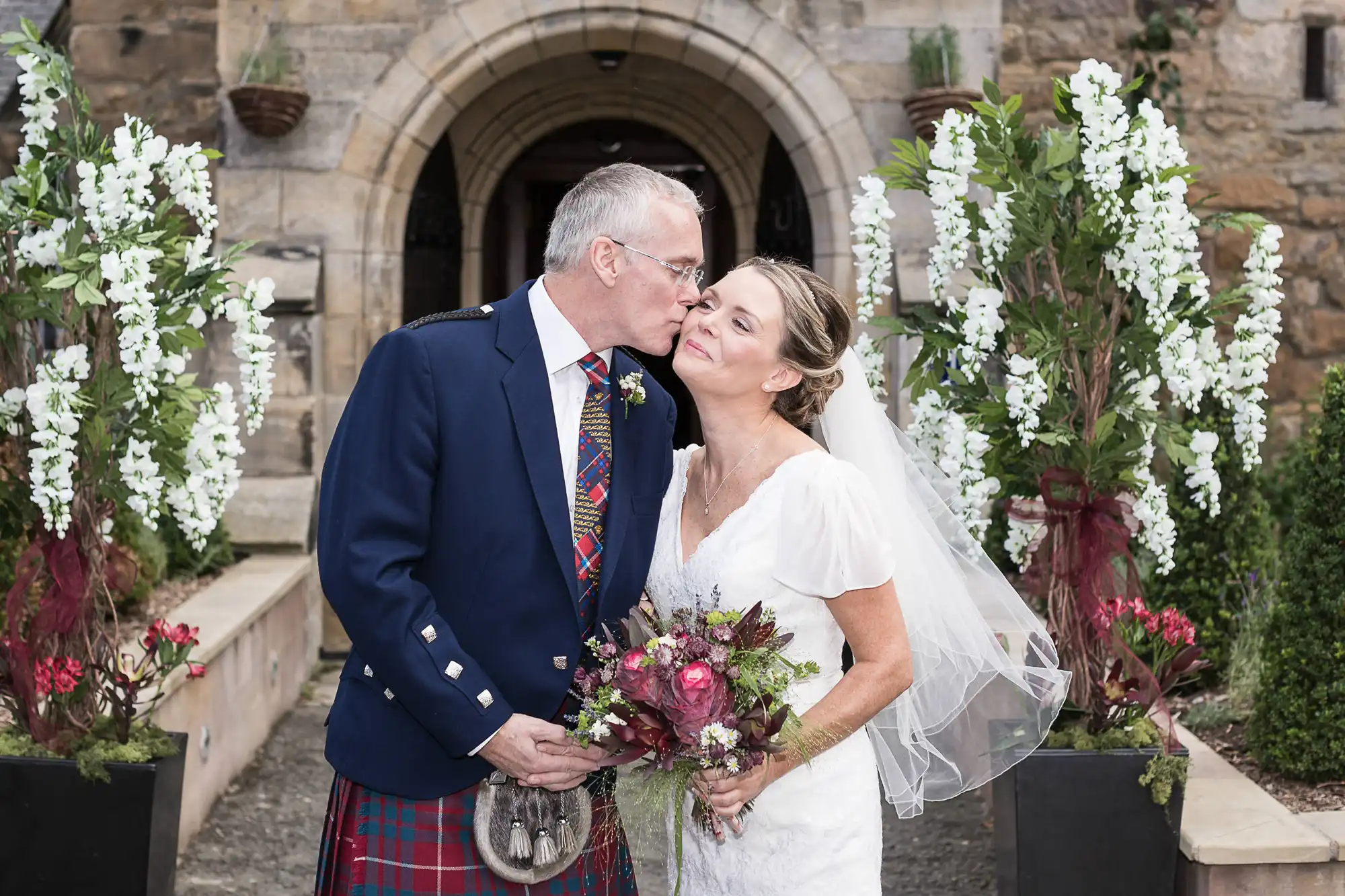 A bride and groom kissing outside a stone church, the groom in a tartan kilt and the bride in a white dress, holding a bouquet, with white floral arrangements on either side.