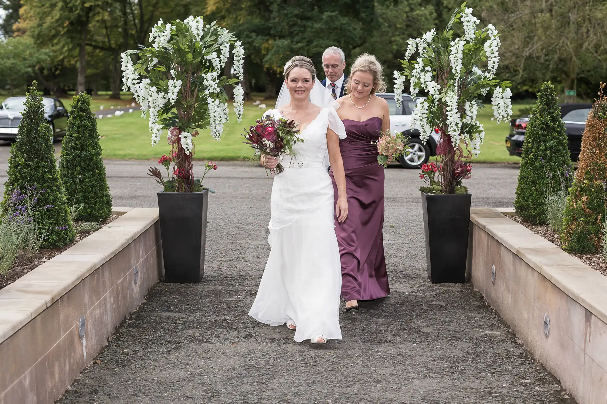 A bride in a white dress and a bridesmaid in a purple gown walking along a garden aisle, with floral arrangements on pedestals and a man in the background.