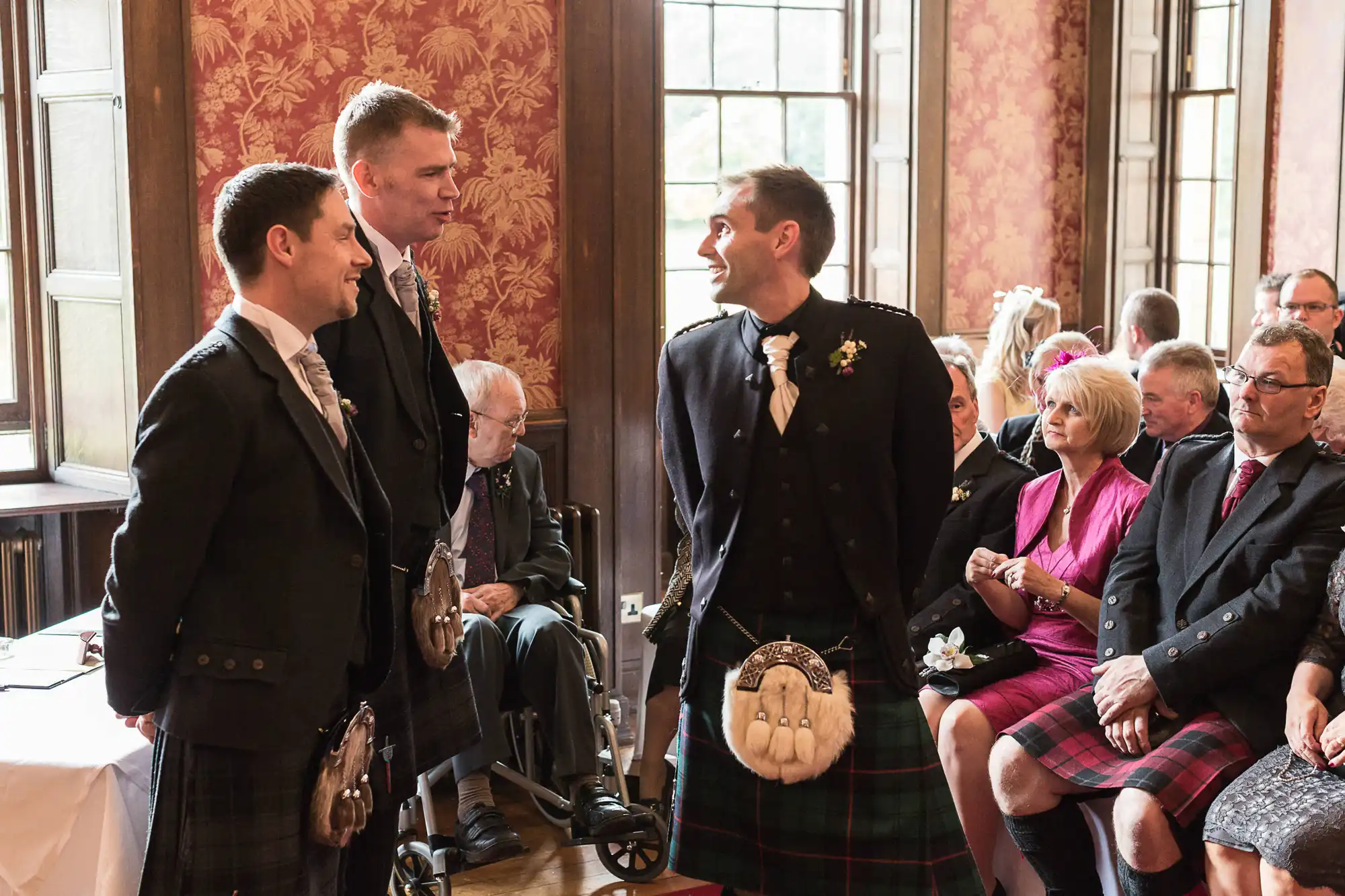 Two men in traditional scottish kilts smiling at each other in a wedding ceremony, with guests seated around in a decorated room.