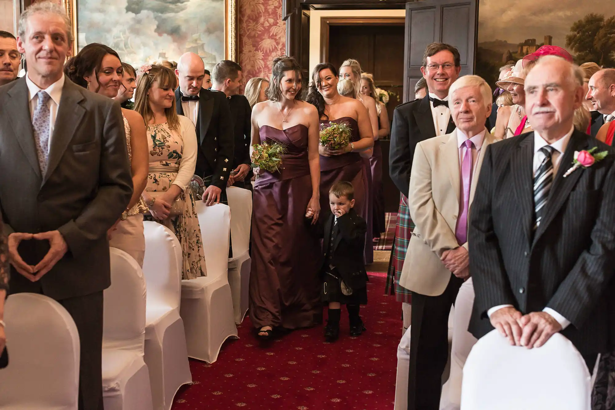 Guests smiling and watching as a bride walks down the aisle at a wedding ceremony, with a young boy in a kilt looking shyly towards the ground.