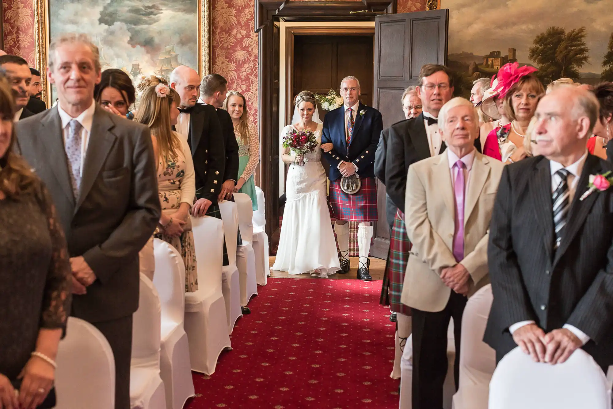 Bride walking down the aisle with her father who wears a kilt, in a room with guests on either side and historical paintings on the walls.