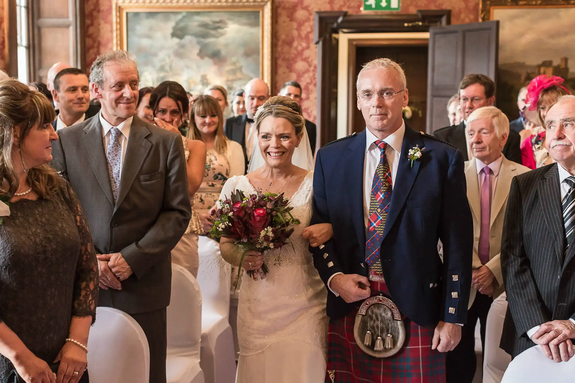 A bride and groom, the groom in a kilt, walk down the aisle smiling, surrounded by guests in a room with ornate decorations and paintings.