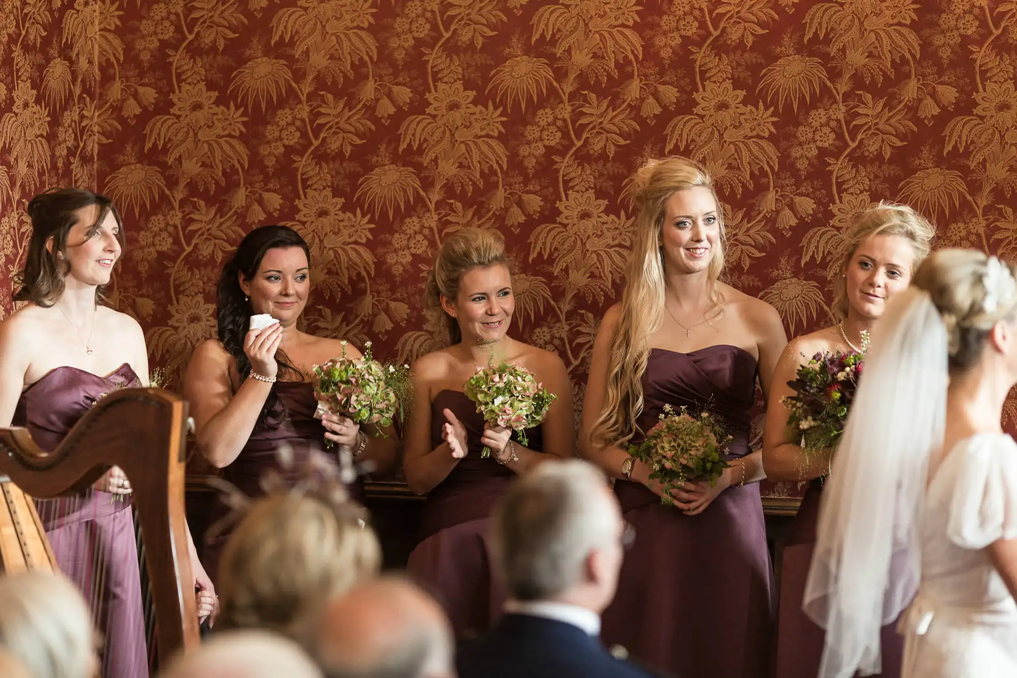 Bridesmaids in plum dresses holding bouquets, watching a bride approach in a ceremony room with ornate golden wallpaper.