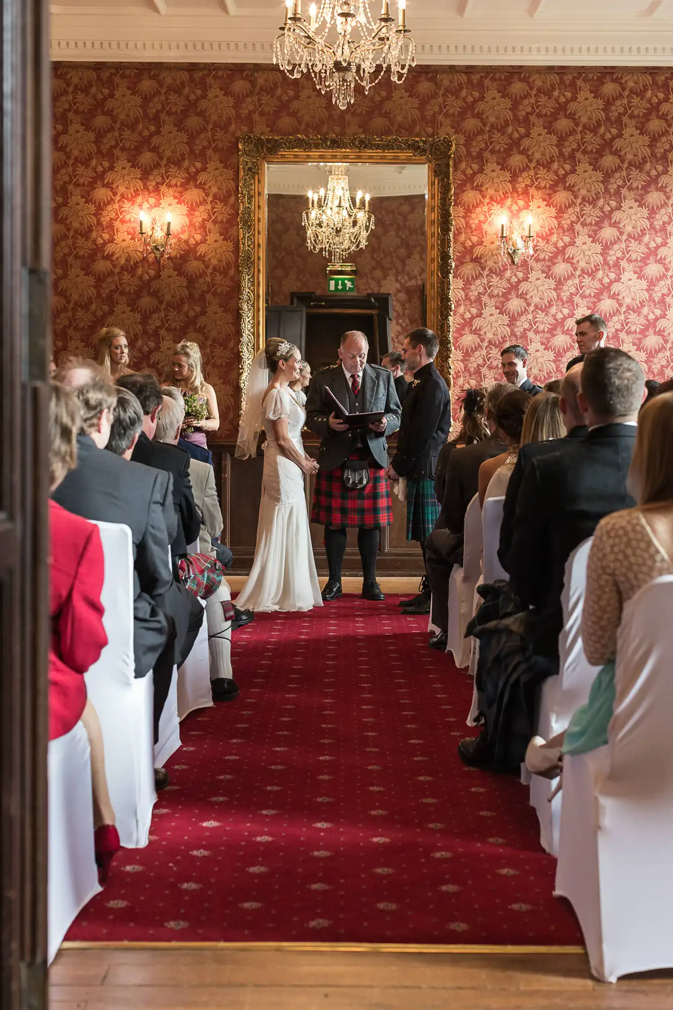 A wedding ceremony in a grand room with red patterned wallpaper, featuring a couple at the altar with a man in a kilt officiating and guests seated along the aisle.