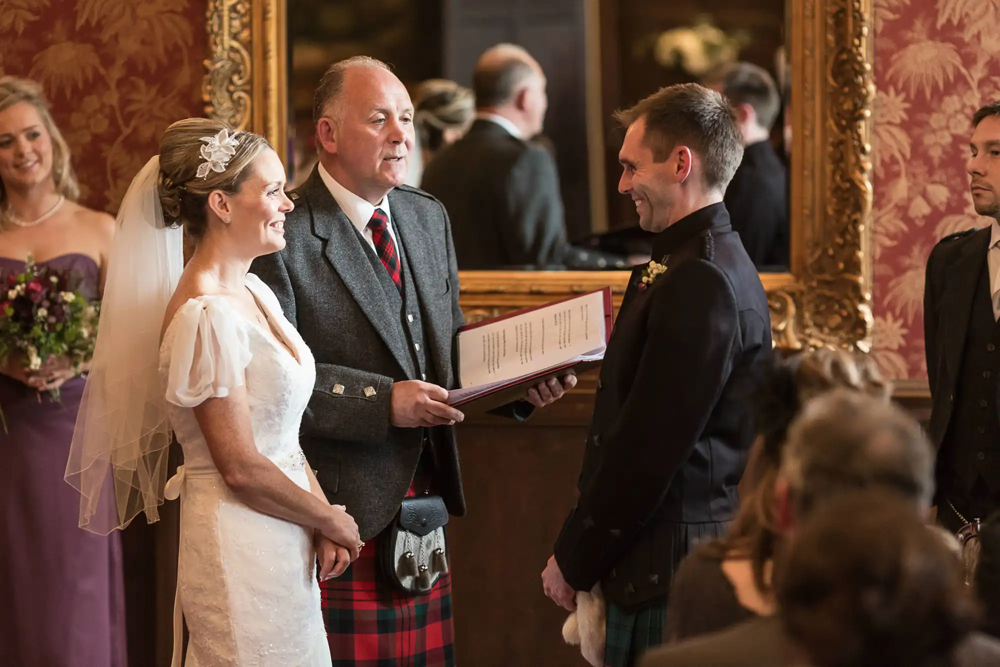 A bride and groom smiling at each other during a wedding ceremony, with an officiant in traditional scottish attire holding a booklet.