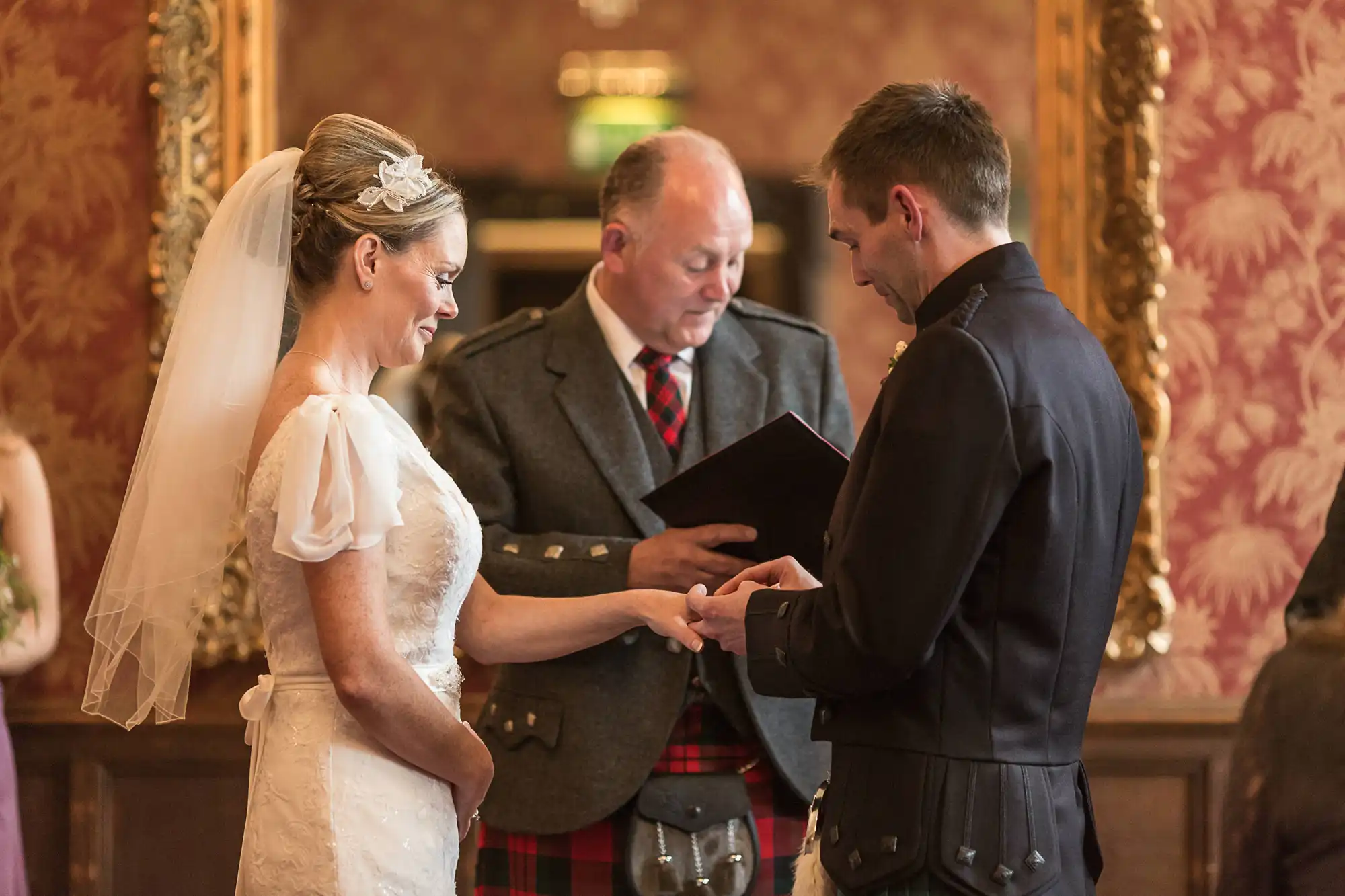 Bride and groom exchange rings during a wedding ceremony, officiated by a man in a kilt, in a richly decorated room.