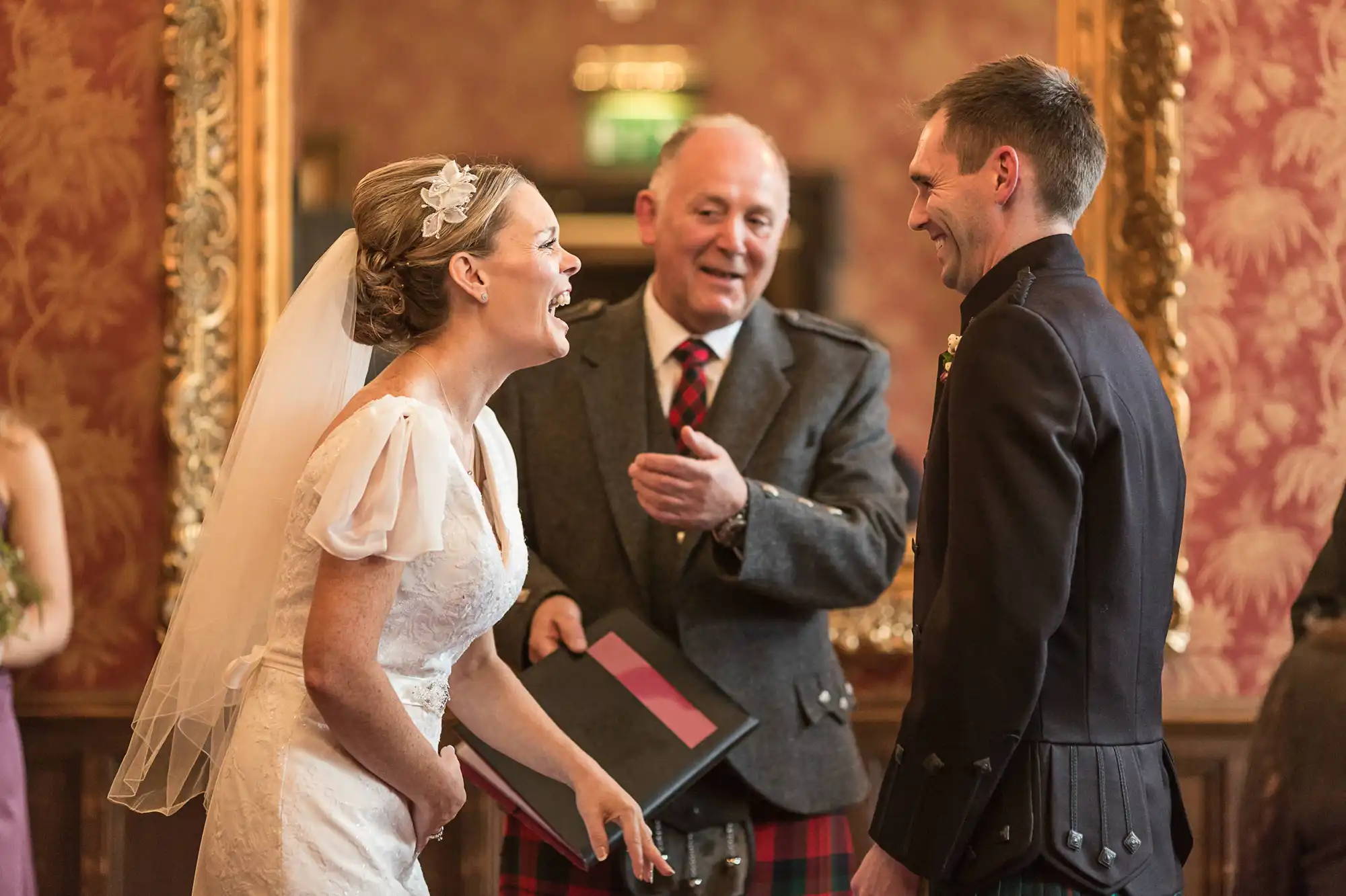 A bride in a white dress and a groom in a military uniform laugh joyfully, conversing with an older man in a kilt at a wedding.