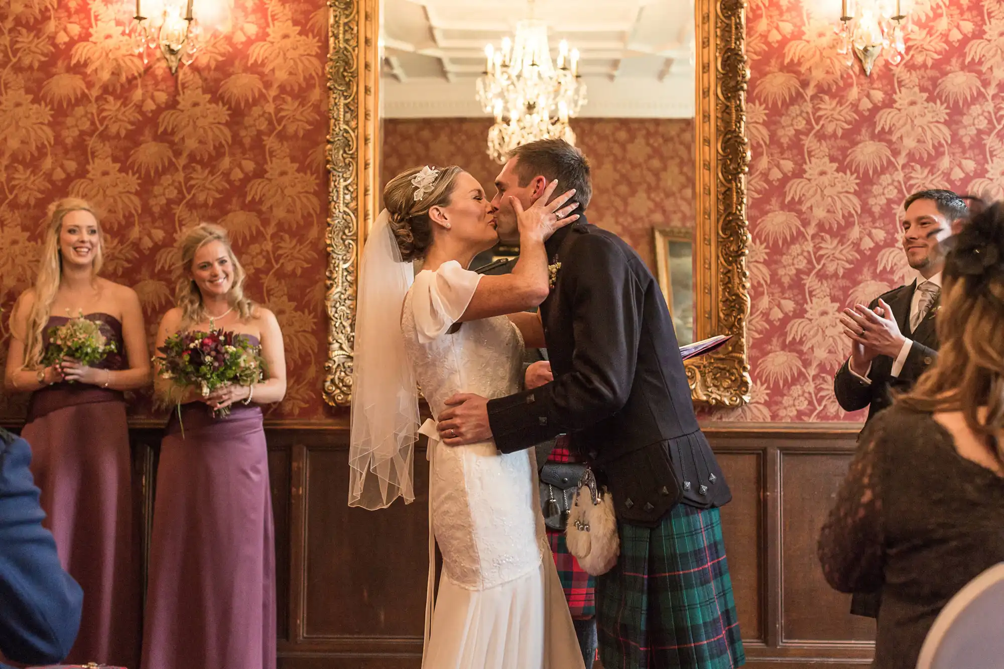 A bride in a white dress and a groom in a kilt kiss passionately in a room with elegantly dressed guests looking on.