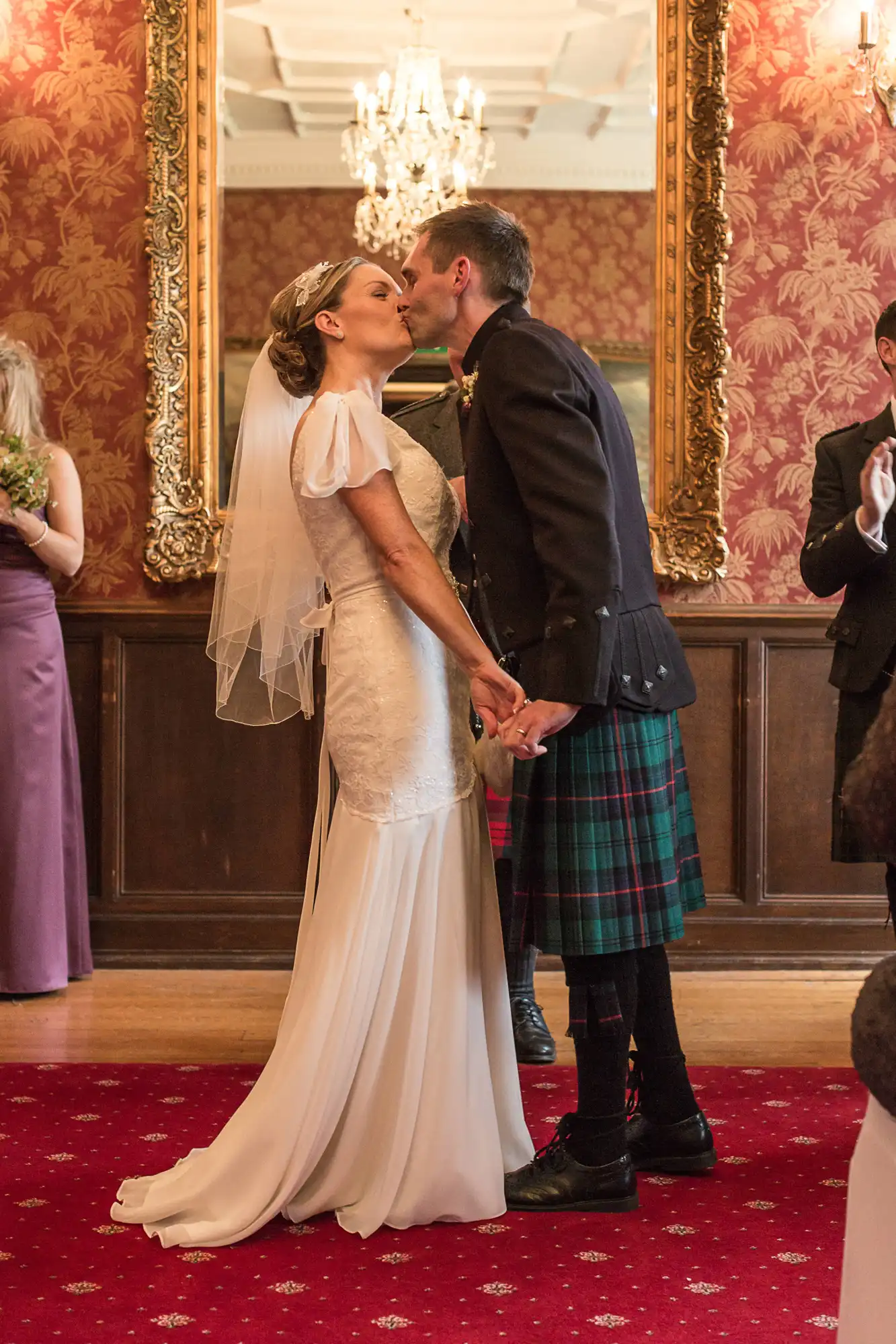 Bride in a white dress and groom in a kilt kissing at their wedding ceremony, with guests and ornate décor in the background.