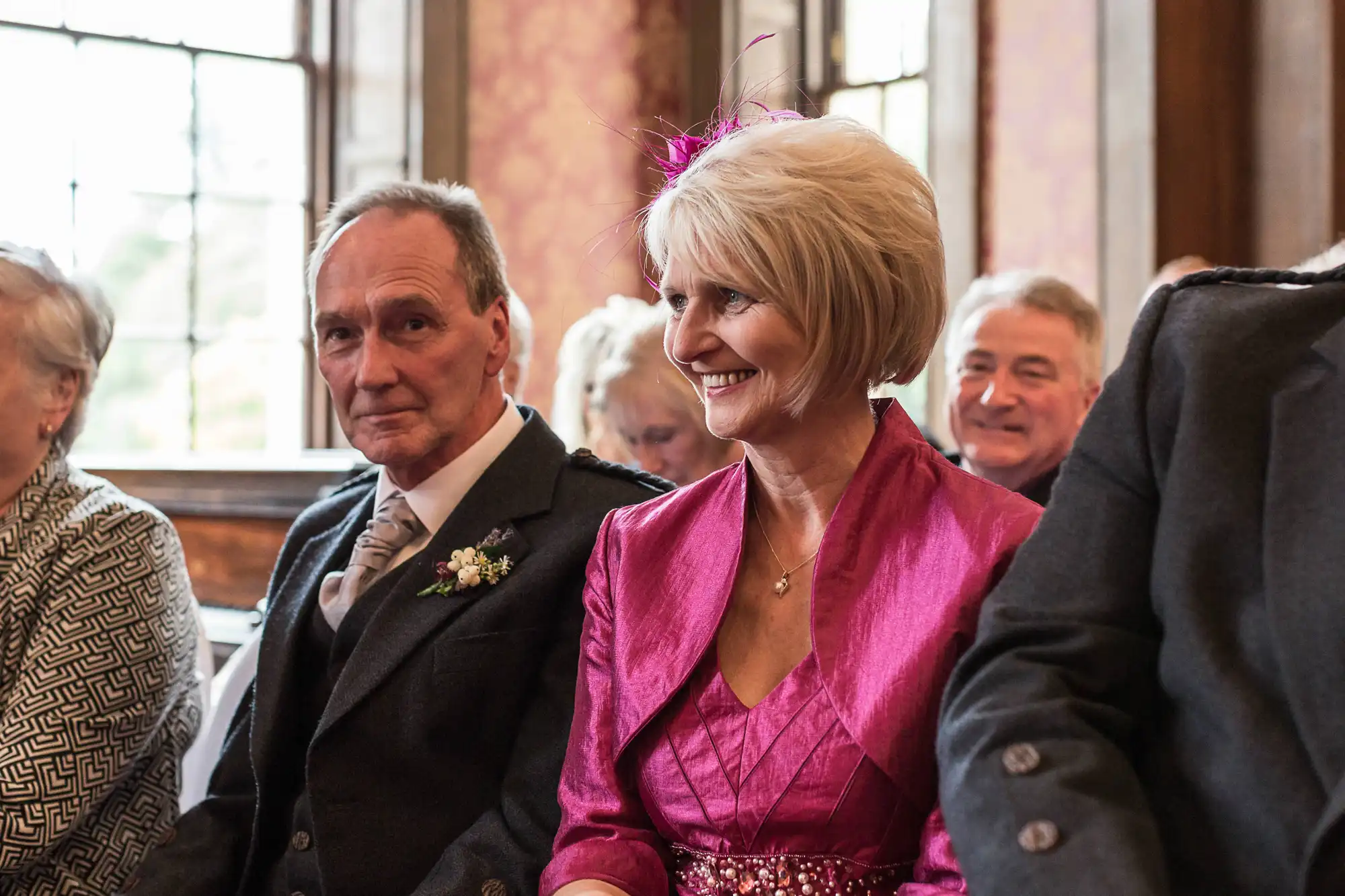 Woman in pink hat and man in suit attending an indoor event, smiling and looking to the side, with other guests in background.