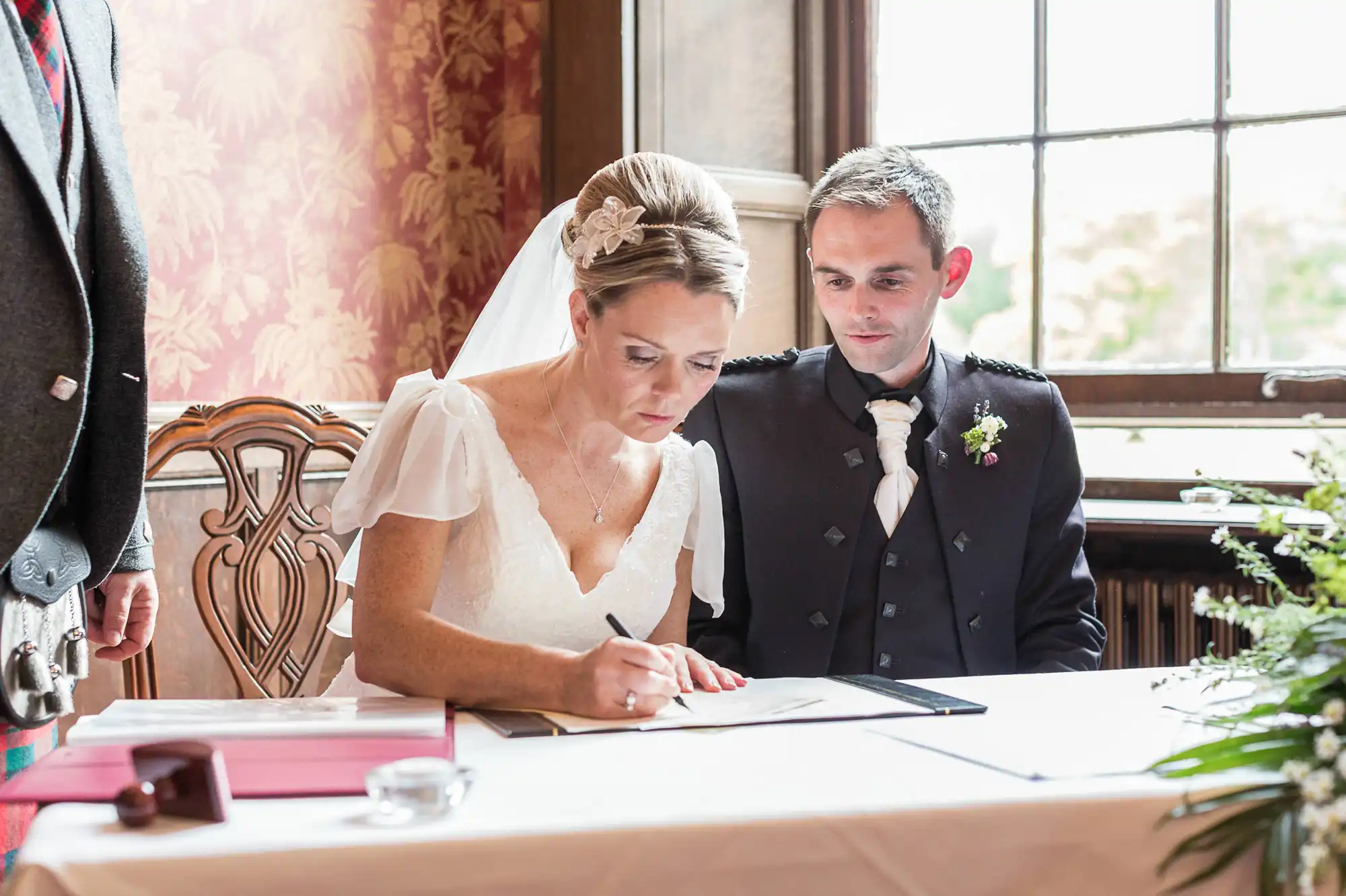 A bride in a white dress signing a document at a table, with the groom in a dark suit watching beside her, indoors by a window.